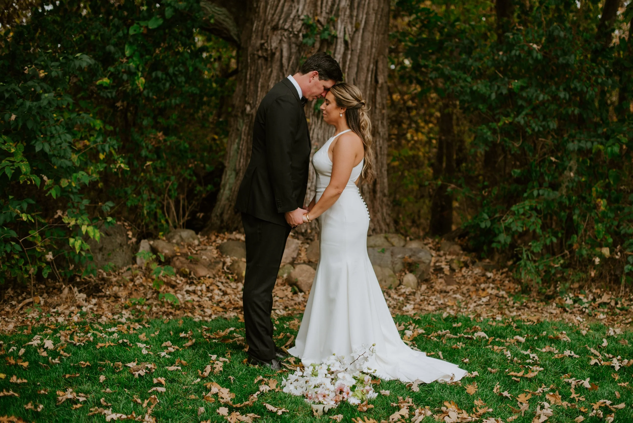 A bride and groom stand close together outdoors, touching foreheads, holding hands, with a large tree in the background. The bride wears a white gown and the groom a black suit, surrounded by fallen leaves on the grass.