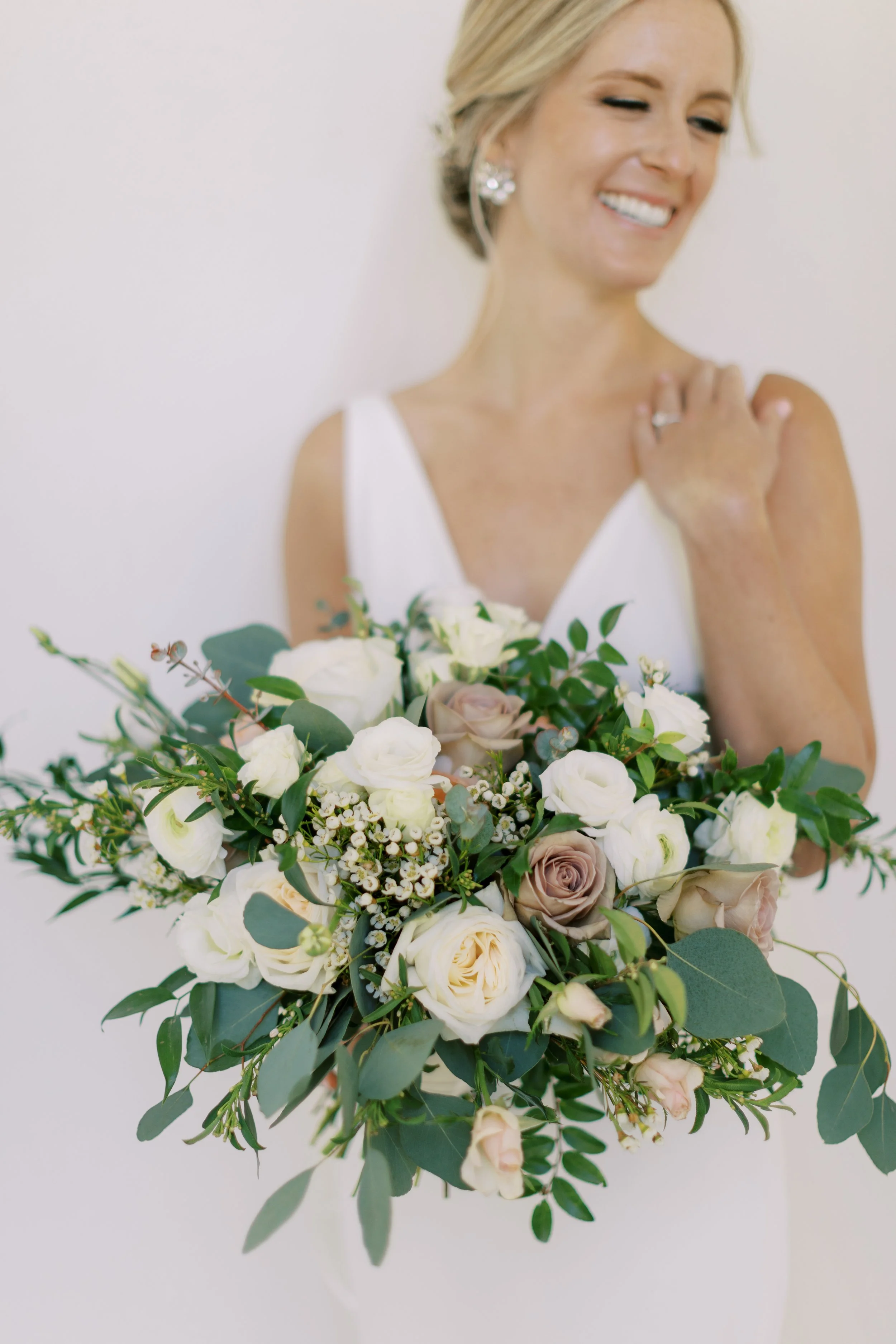 A bride holding a bouquet of white and blush roses with greenery, smiling and looking to the side, against a plain white background.