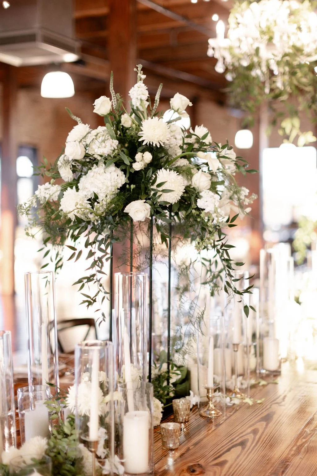 A tall floral centerpiece with white roses, chrysanthemums, and greenery on a wooden table, surrounded by candles and glass vases, in a decorated indoor venue.