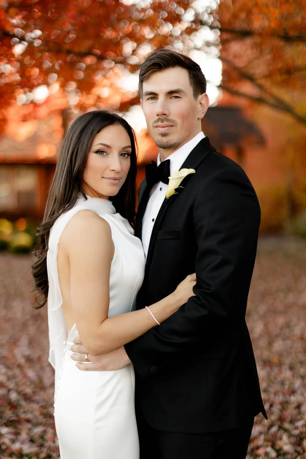 A bride and groom in wedding attire standing outdoors during autumn, with a background of orange and red trees.