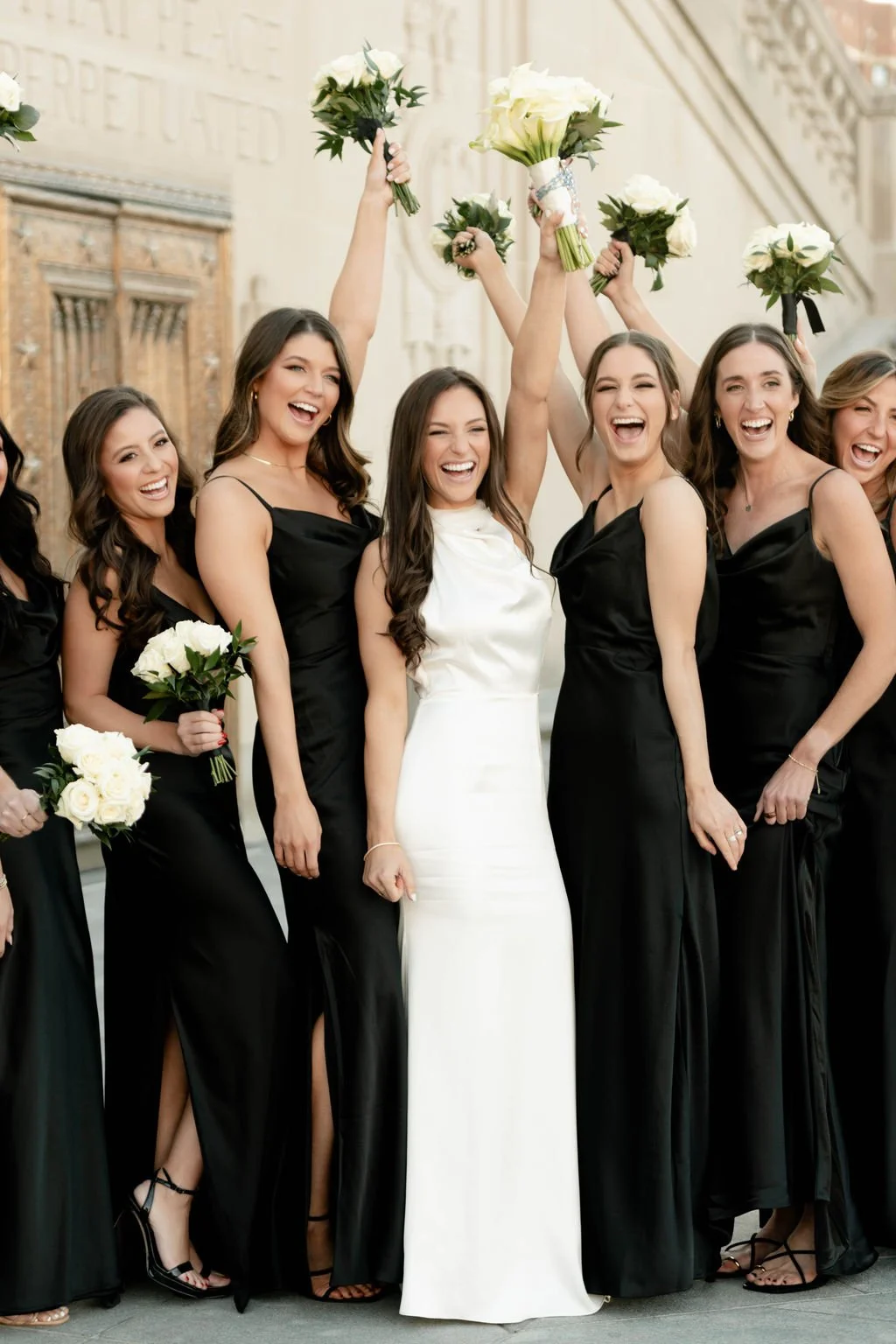 Group of women in black dresses and one woman in a white dress celebrating with bouquets of white roses outside a building.