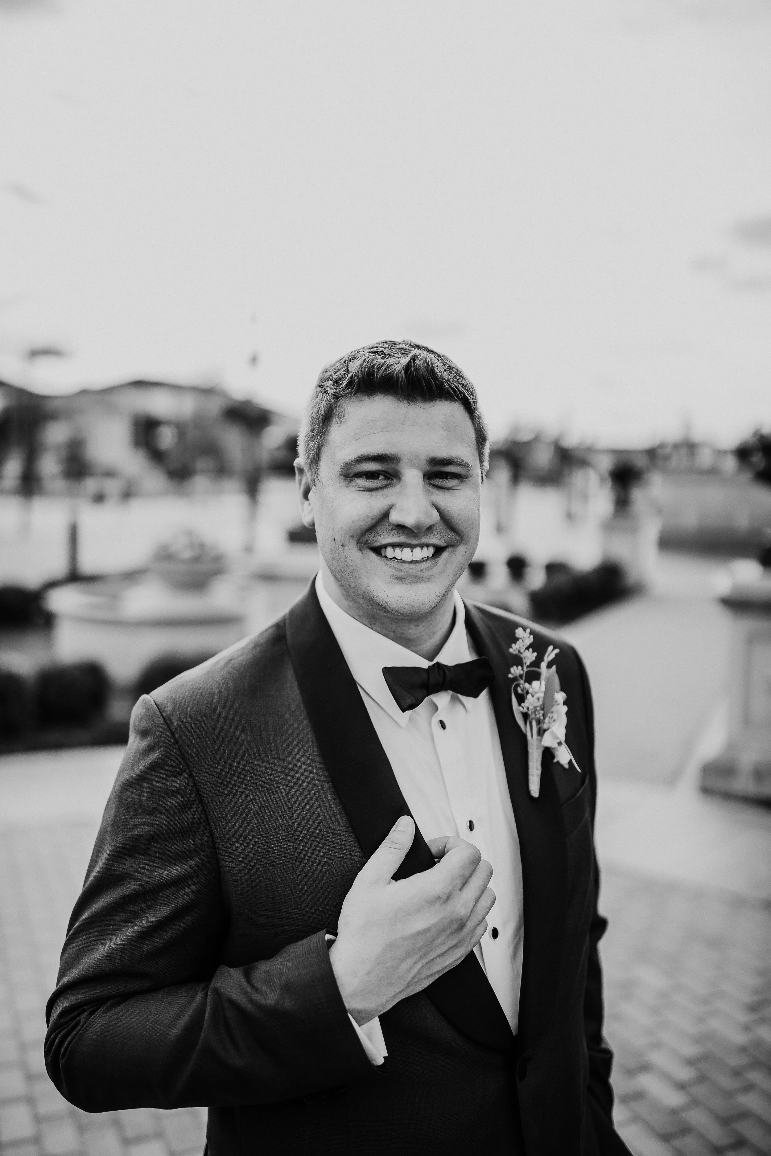 A smiling man dressed in a tuxedo with a bow tie and boutonniere, standing outdoors with blurred buildings and a fountain in the background.