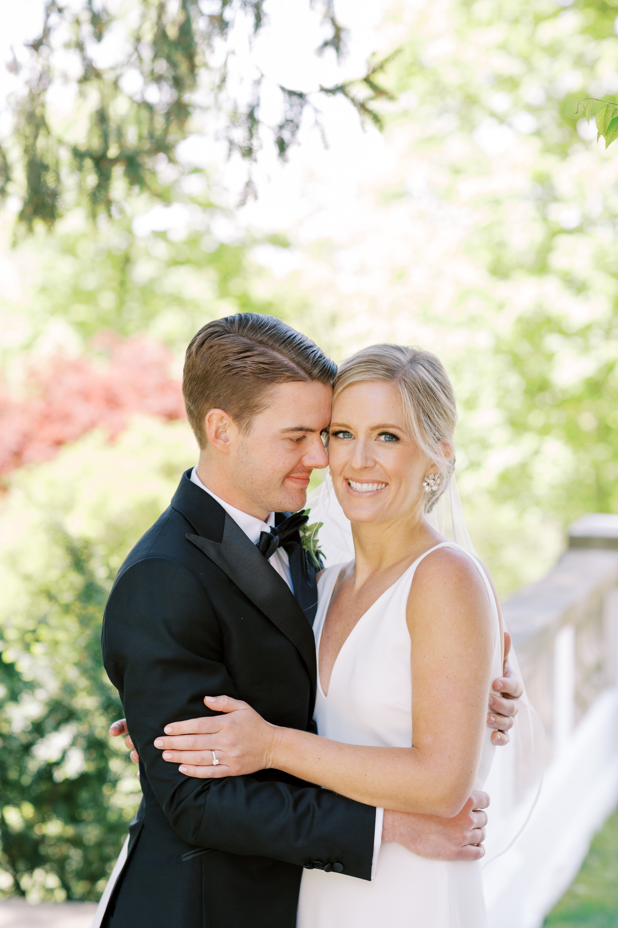 A bride and groom are embracing outdoors in a park with green trees. The groom is wearing a black tuxedo and the bride is in a white wedding dress. They are smiling and close together, with the groom whispering in the bride's ear.