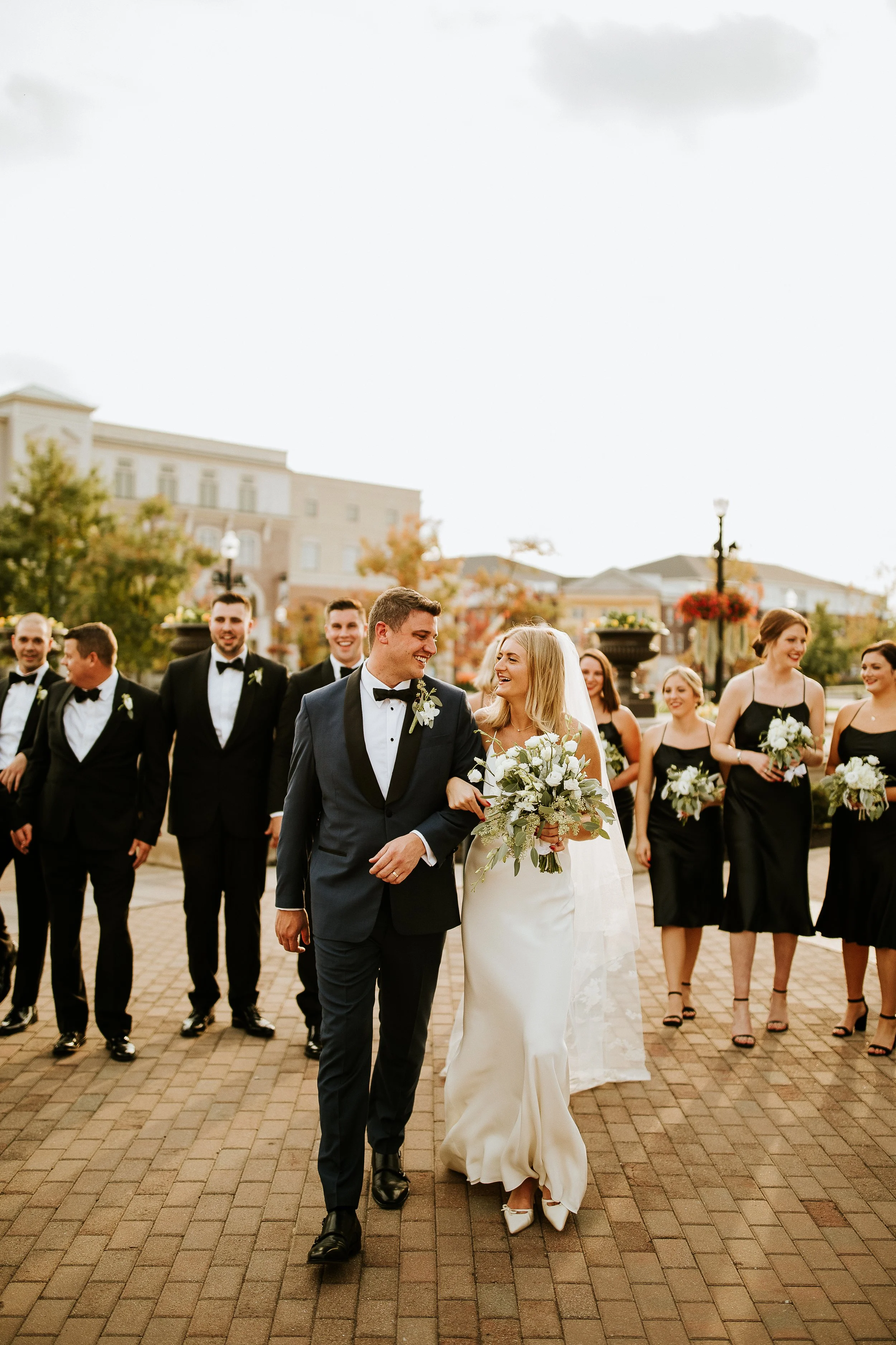 Wedding party walking outdoors on a brick path with buildings and autumn trees in the background. The bride and groom are in the foreground, smiling and holding hands, surrounded by bridesmaids and groomsmen dressed in formal attire.