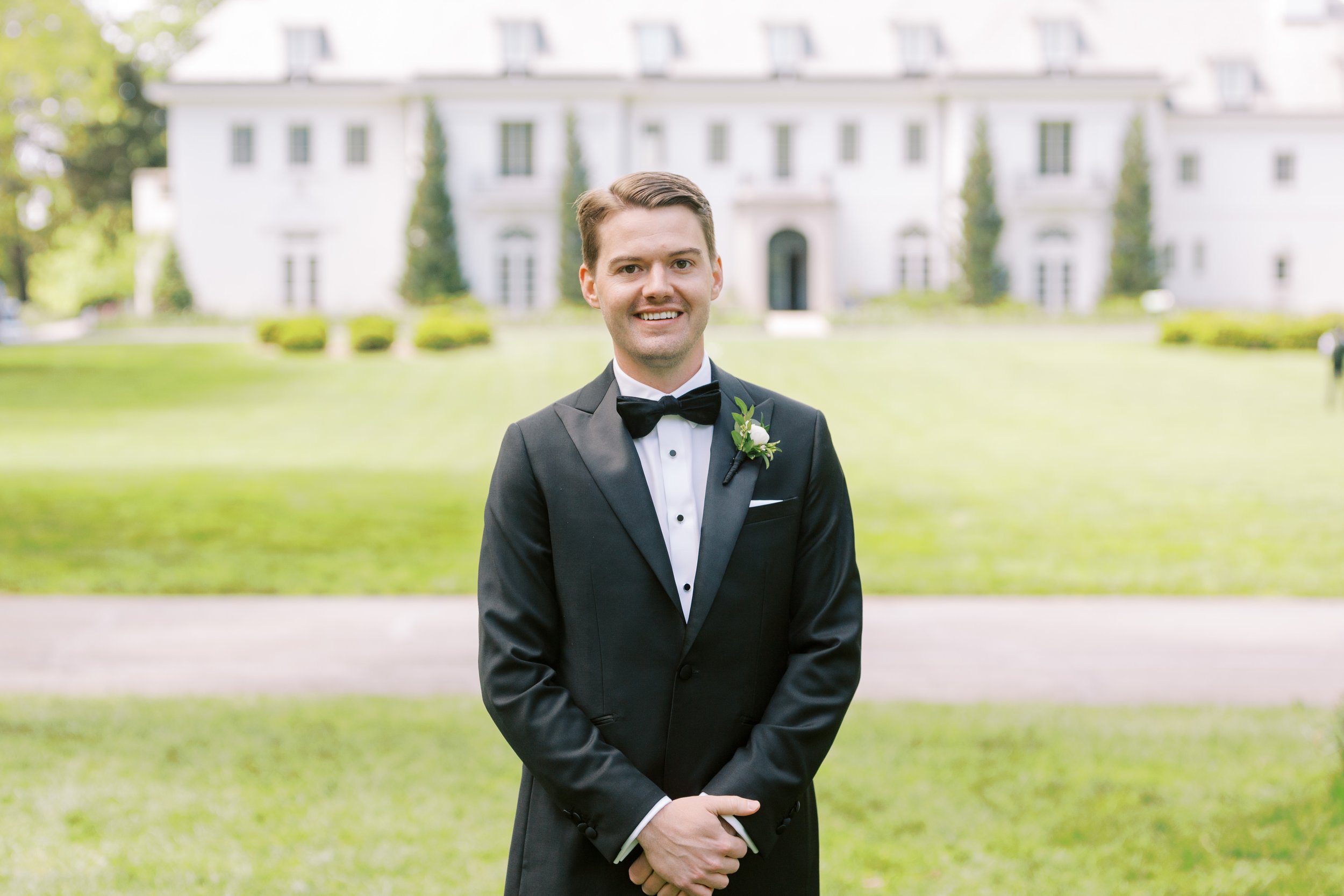 Portrait of a young man in a tuxedo with a boutonniere, standing outdoors in front of a large, white house with manicured lawns and tall trees.