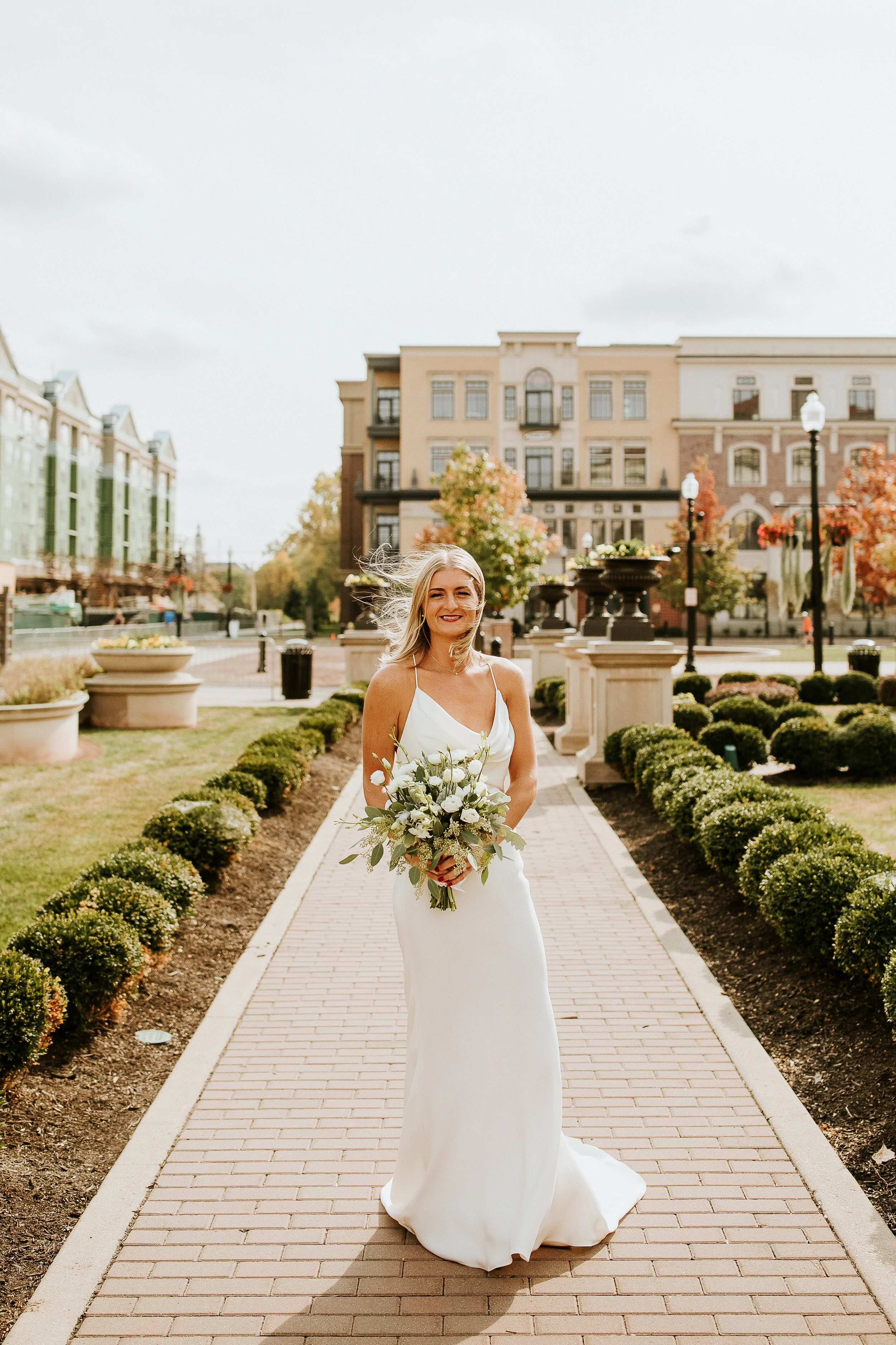A bride in a white wedding dress holding a bouquet of flowers, standing on a brick pathway in a landscaped outdoor area with buildings and autumn trees in the background.