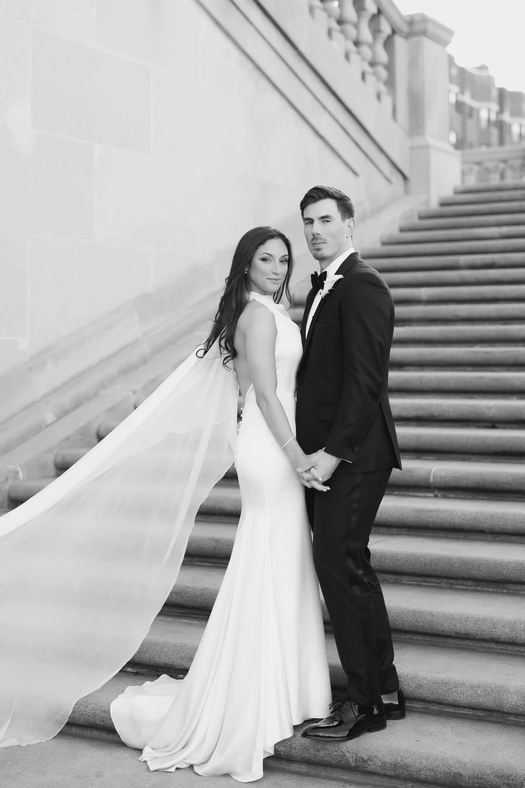 A bride and groom holding hands on outdoor staircase, dressed in wedding attire, with the bride in a gown and veil and the groom in a tuxedo, in black and white.