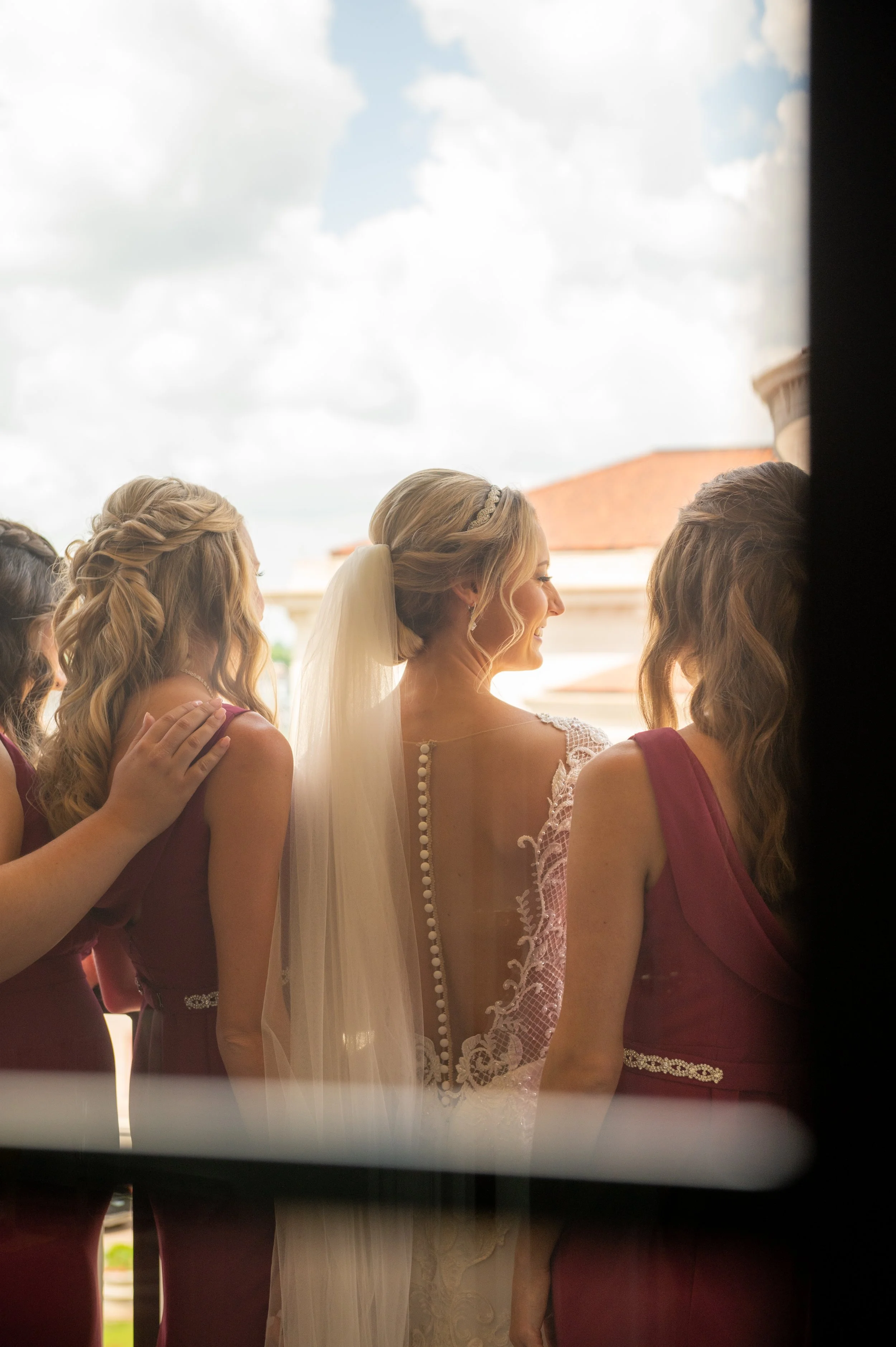 A group of women dressed in formal attire, with a bride wearing a veil in the center, seen through a narrow opening with a cloudy sky in the background.