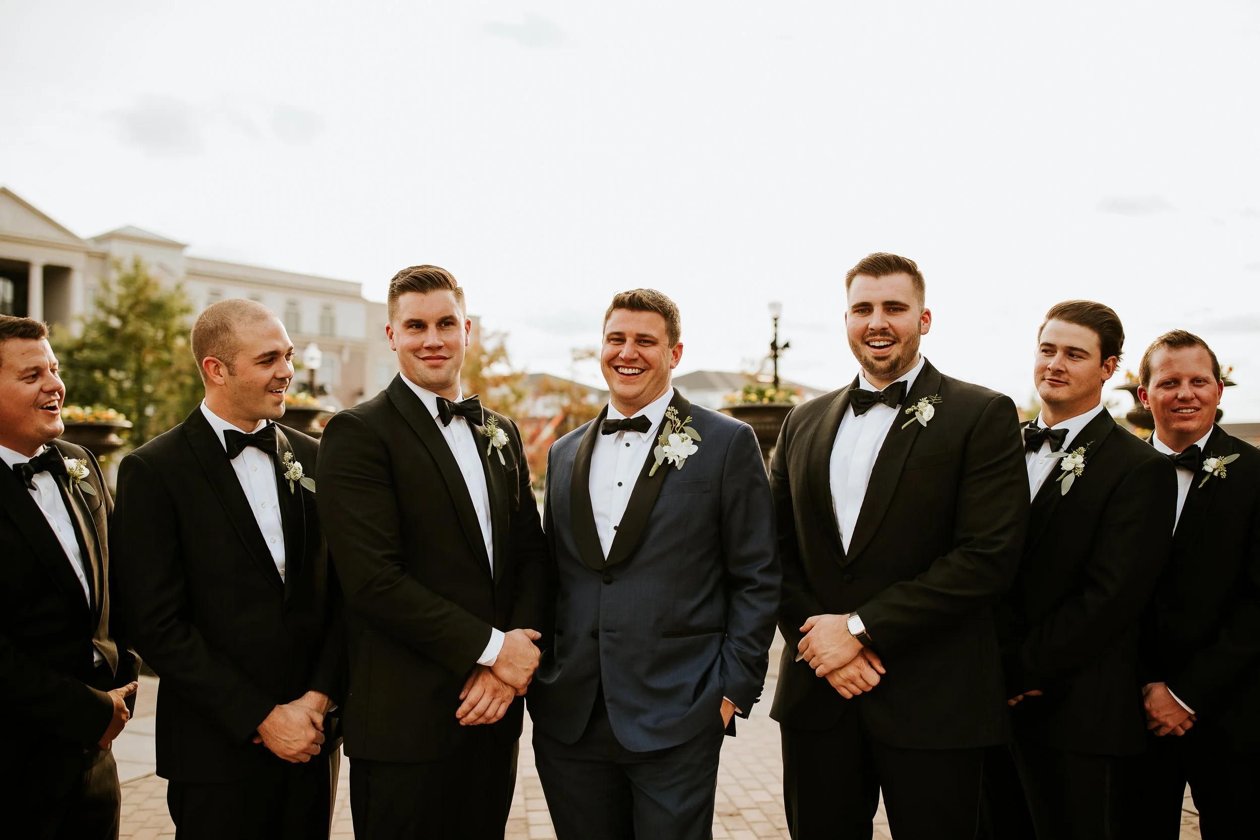 Group of men in formal tuxedos, standing outdoors, smiling and talking to each other.