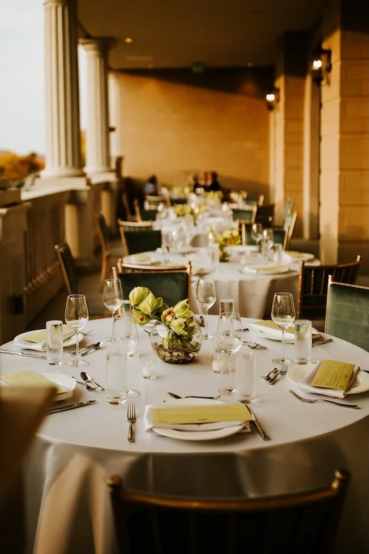 Elegantly set banquet table with white tablecloth, floral centerpiece, wine glasses, water glasses, plates, and cutlery in a decoratively lit restaurant or event space.