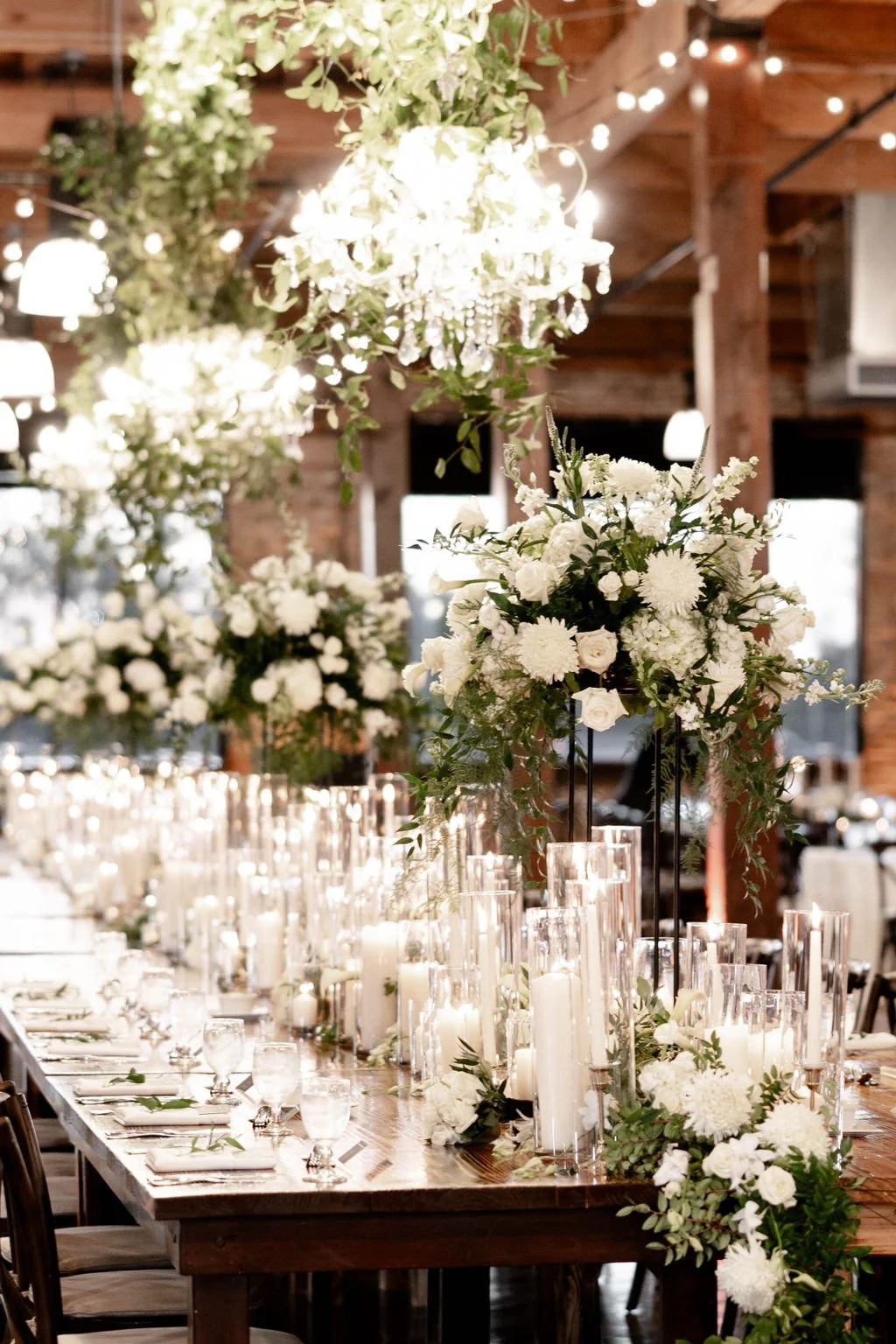 Elegant wedding reception table decorated with tall floral arrangements of white roses, hydrangeas, and greenery, surrounded by numerous white candles in glass holders, with a rustic wooden interior and string lights overhead.