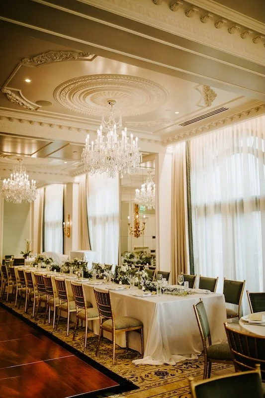 Elegant banquet room with long table set for a formal event, chandeliers hanging from ornate ceiling, large windows with curtains, and decorative wall sconces.
