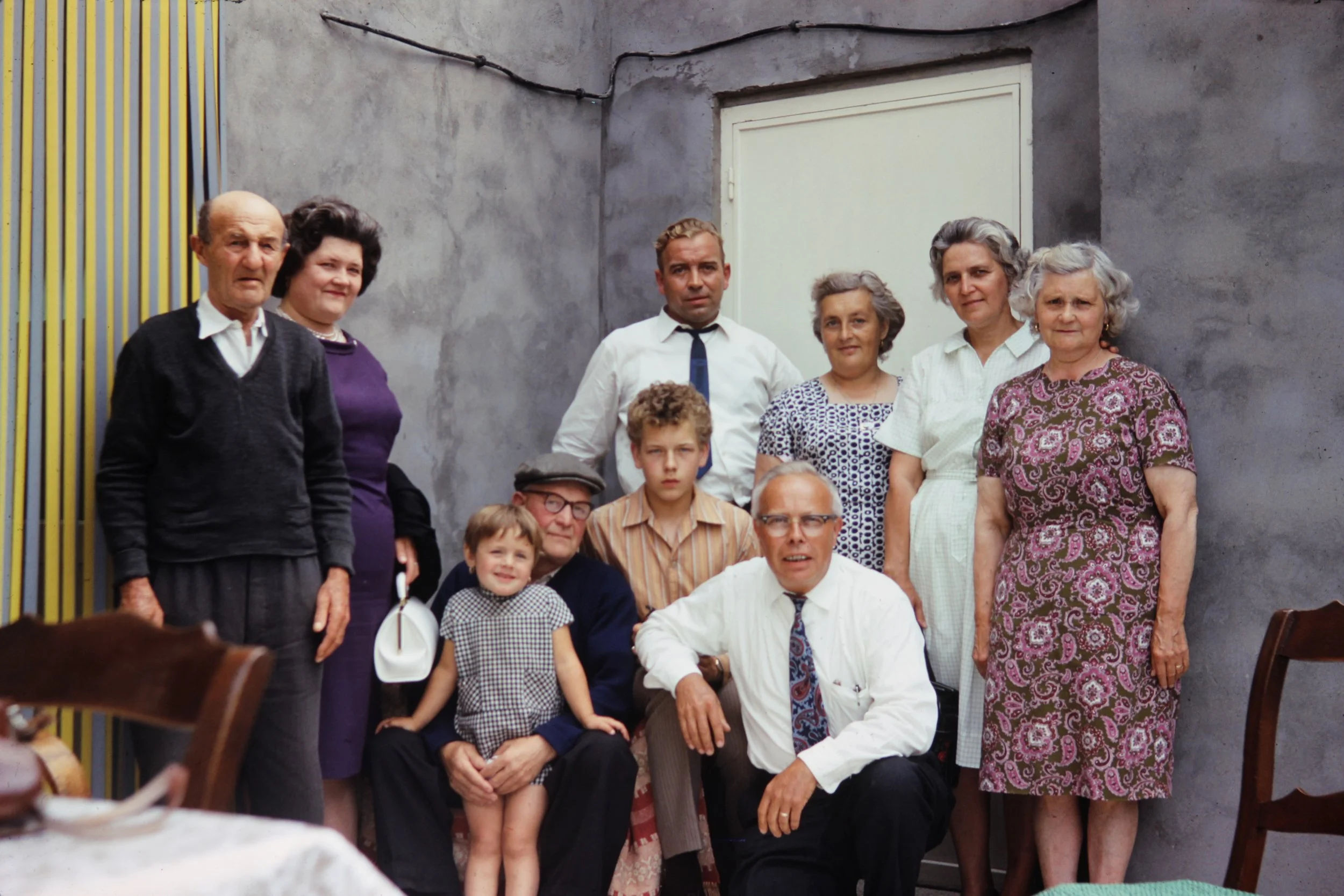 A family photo of twelve people, including children and adults, gathered indoors against a gray wall, dressed in vintage clothing from the mid-20th century.