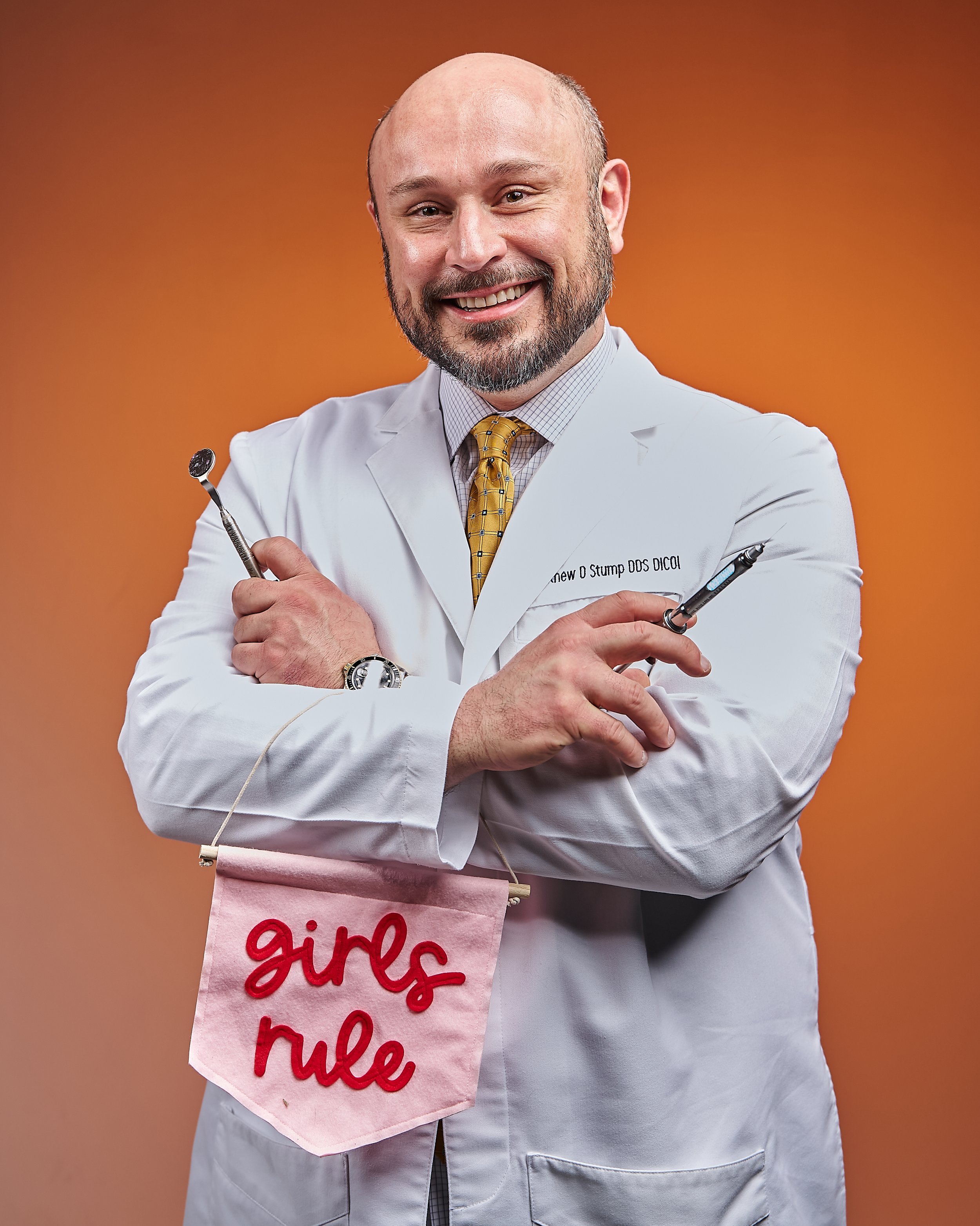 A smiling man in a white lab coat holds dental tools and a small pink banner that reads "girls rule" against an orange background.