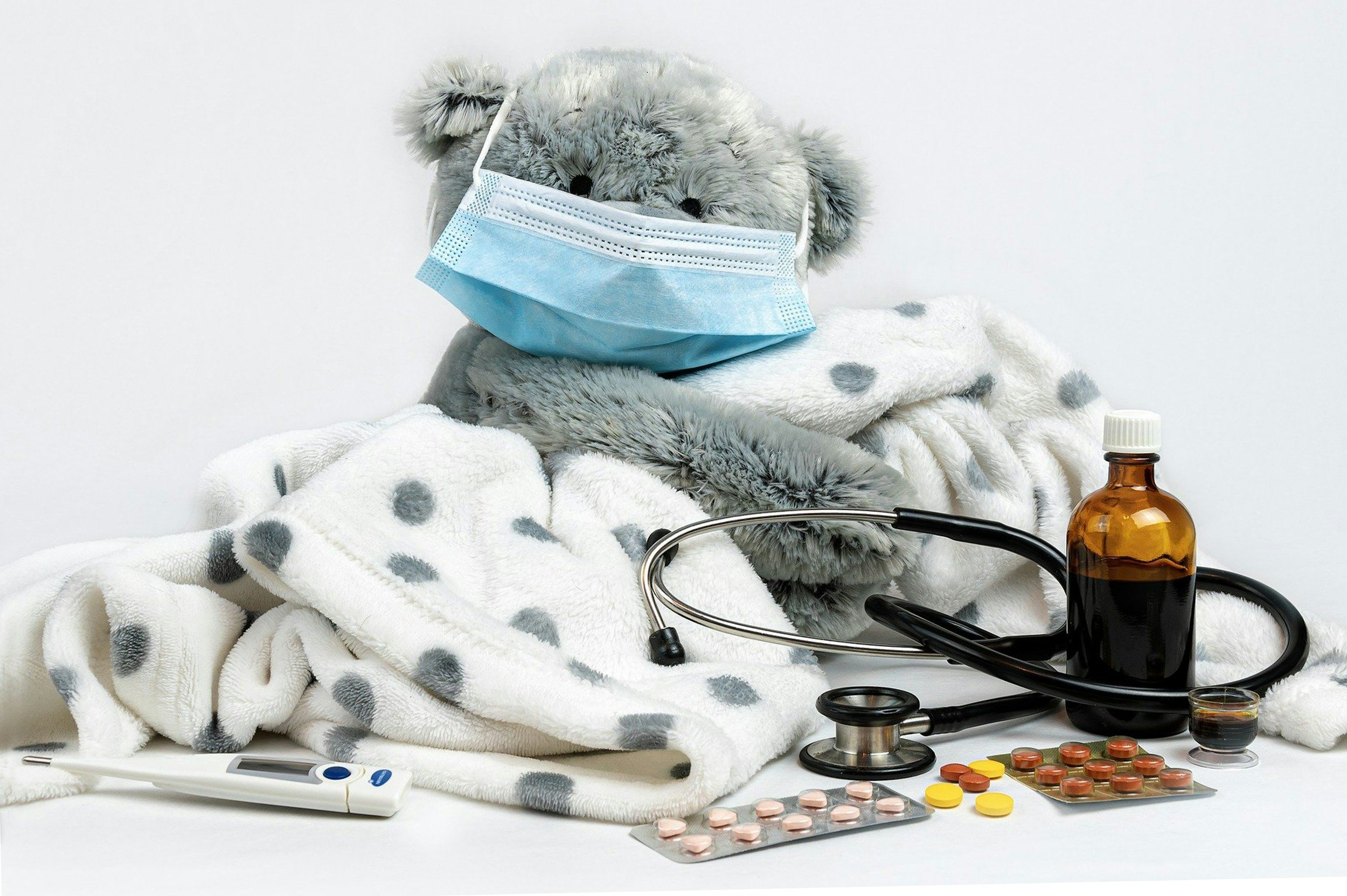 Teddy bear with a face mask, wrapped in a blanket, surrounded by a stethoscope, thermometer, medicine bottles, and blister packs of pills.