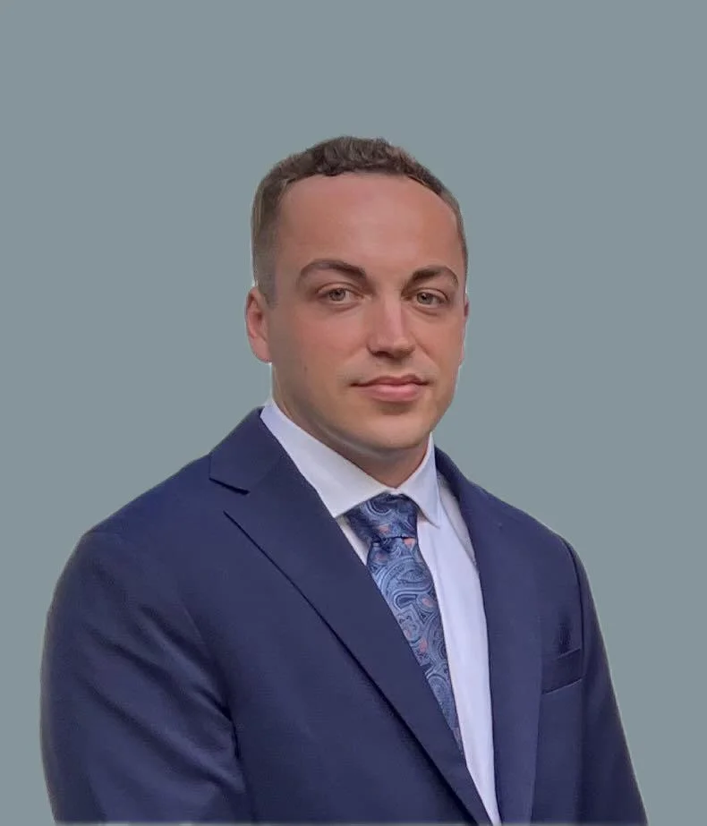 Young man wearing a navy suit, white shirt, and patterned tie, posing against a plain, light blue background.
