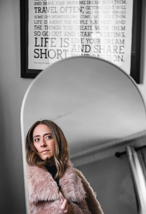 Woman in a pink faux fur coat standing beside an arched mirror with a motivational typographic poster in the background.