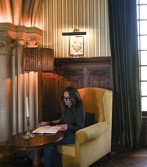 A woman sitting in a yellow armchair reading a book in a warmly lit, elegant room with striped wallpaper, wood paneling, and a candle on a side table