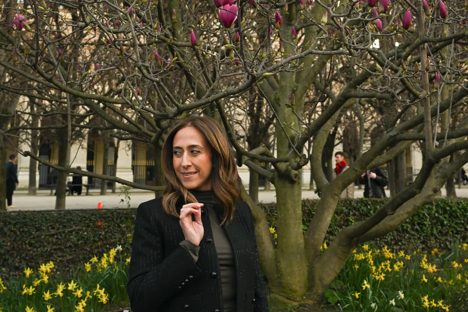 A woman stands in a garden beneath a flowering tree with pink blossoms, with yellow daffodils and a park walkway in the background.