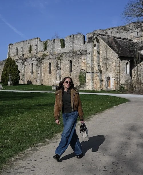 Woman walking along a path in front of ancient stone ruins on a sunny day, wearing a brown shearling jacket, wide-leg jeans, and tinted sunglasses, carrying a white handbag.