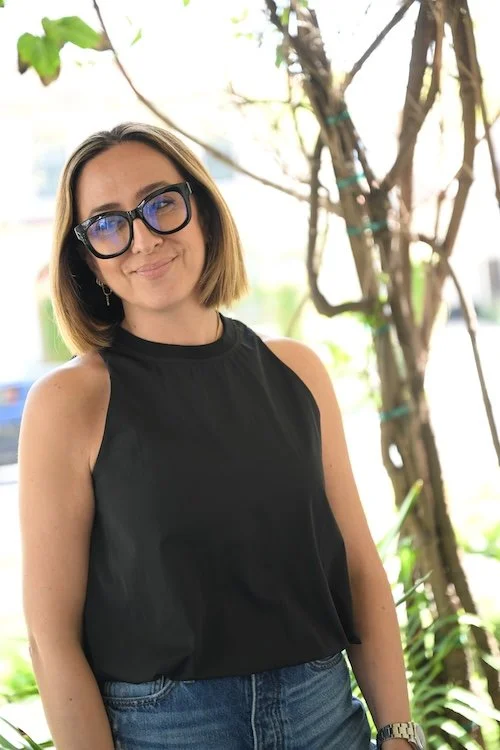 Woman wearing black sleeveless top and glasses, standing outdoors near greenery, smiling softly at the camera.