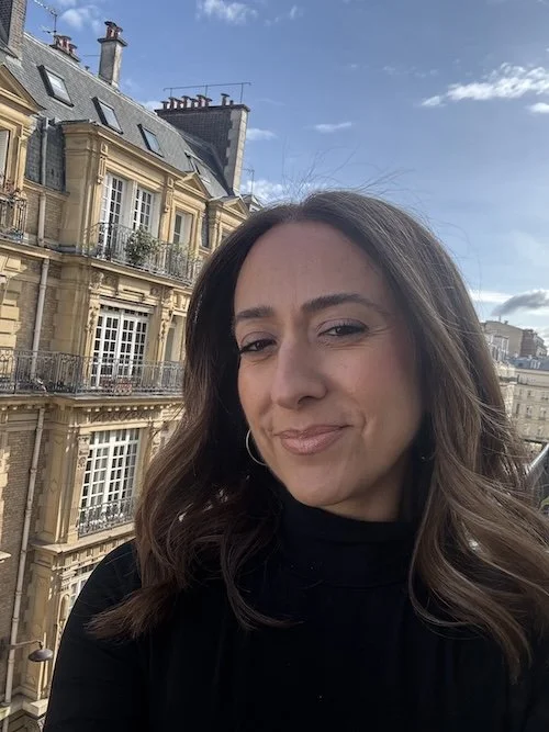 Woman smiling outdoors with a Parisian building in the background.