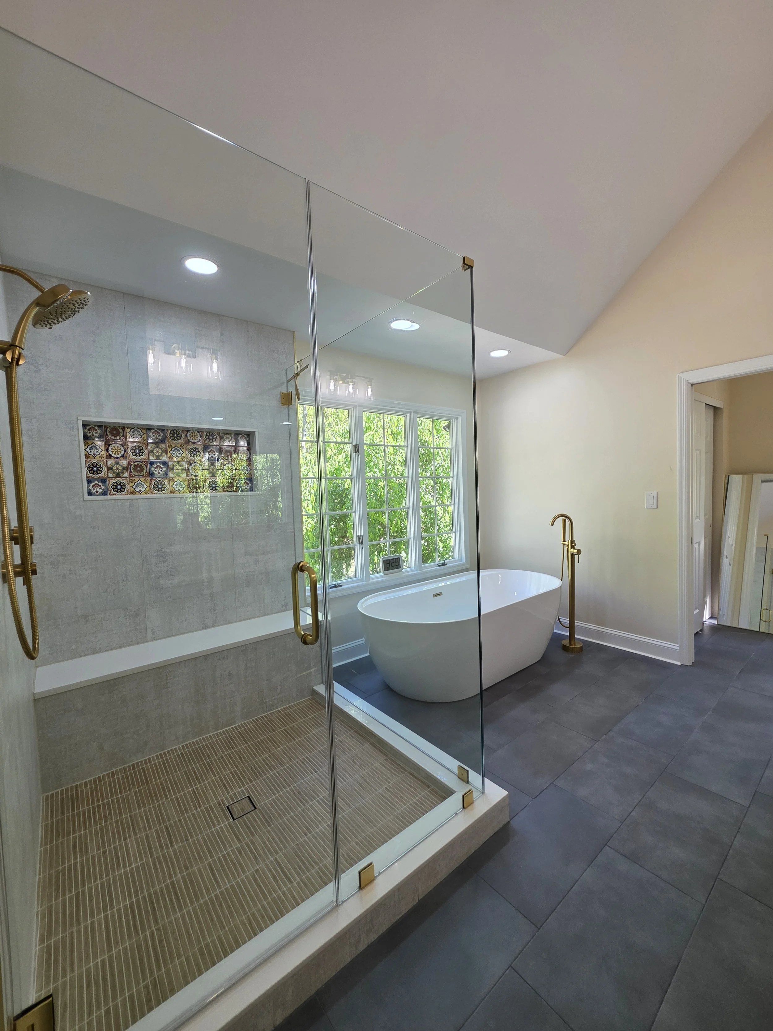 Glass shower enclosure beside the freestanding tub with gold-tone fixtures and large wall tiles.