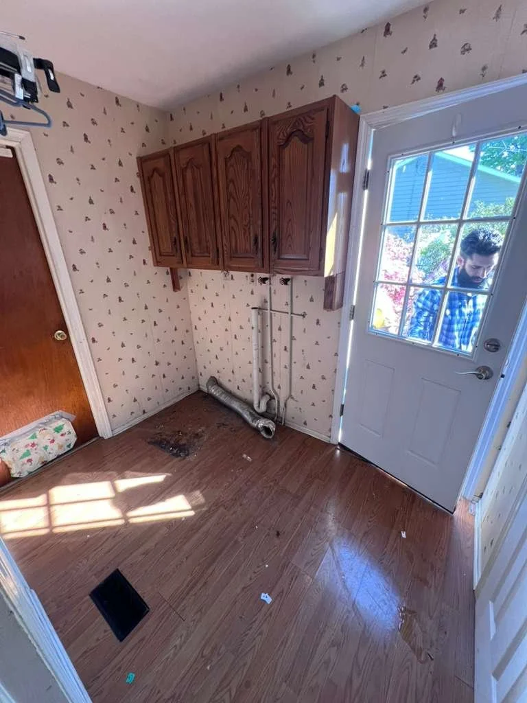 Before renovation: the laundry room with wallpaper, older wall cabinets and exposed appliance connections.