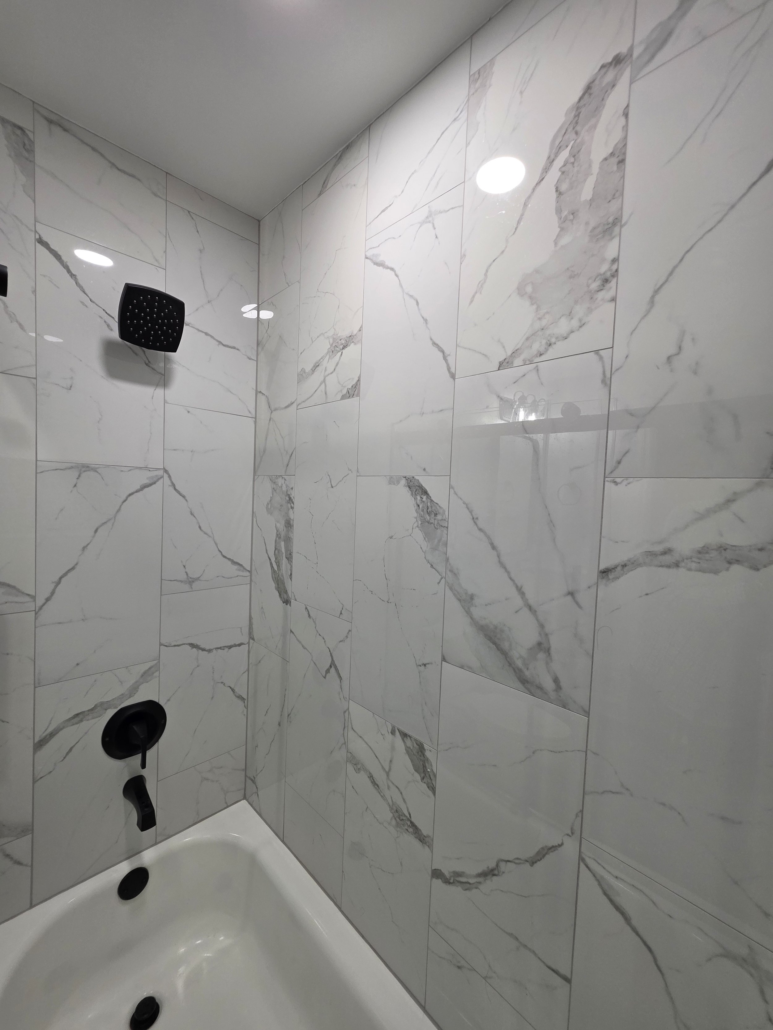 Black shower fixtures above the white tub against marble-look tile.