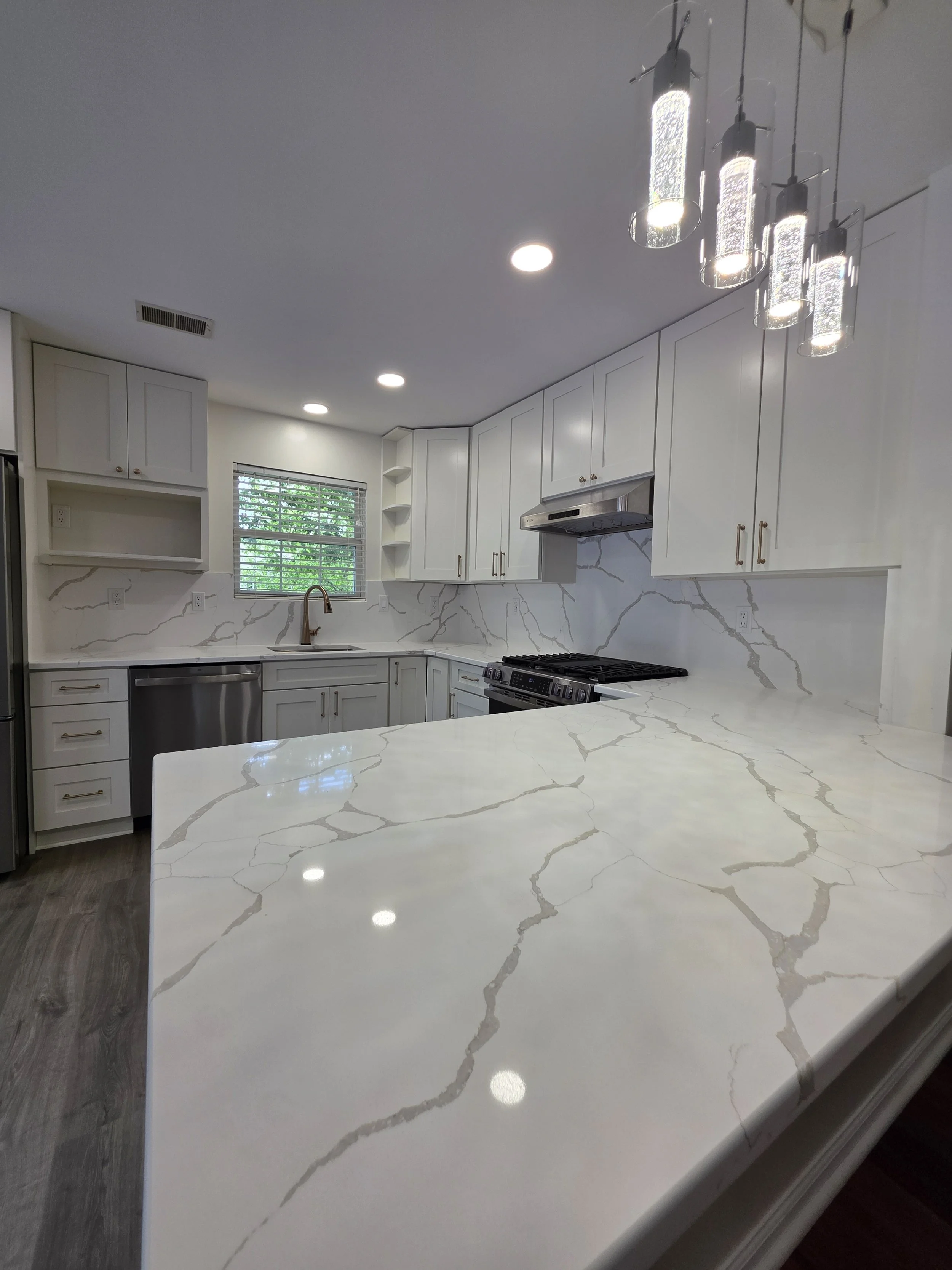 Veined peninsula countertop beneath pendant lights, looking toward the white kitchen cabinets.