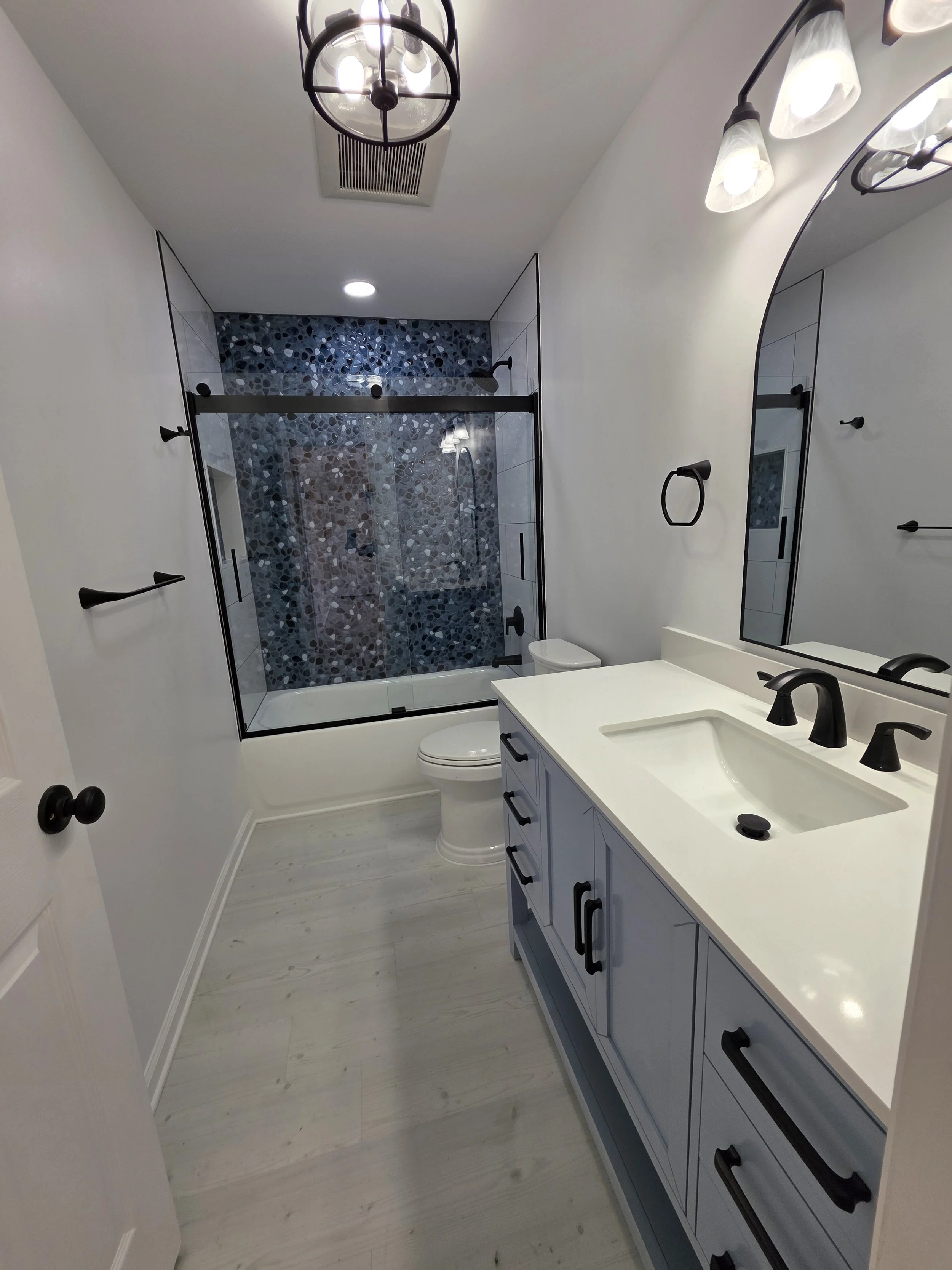 Wide view from the doorway of Lorraine and Ray’s remodeled hallway bathroom, with gray flooring and black fixtures.