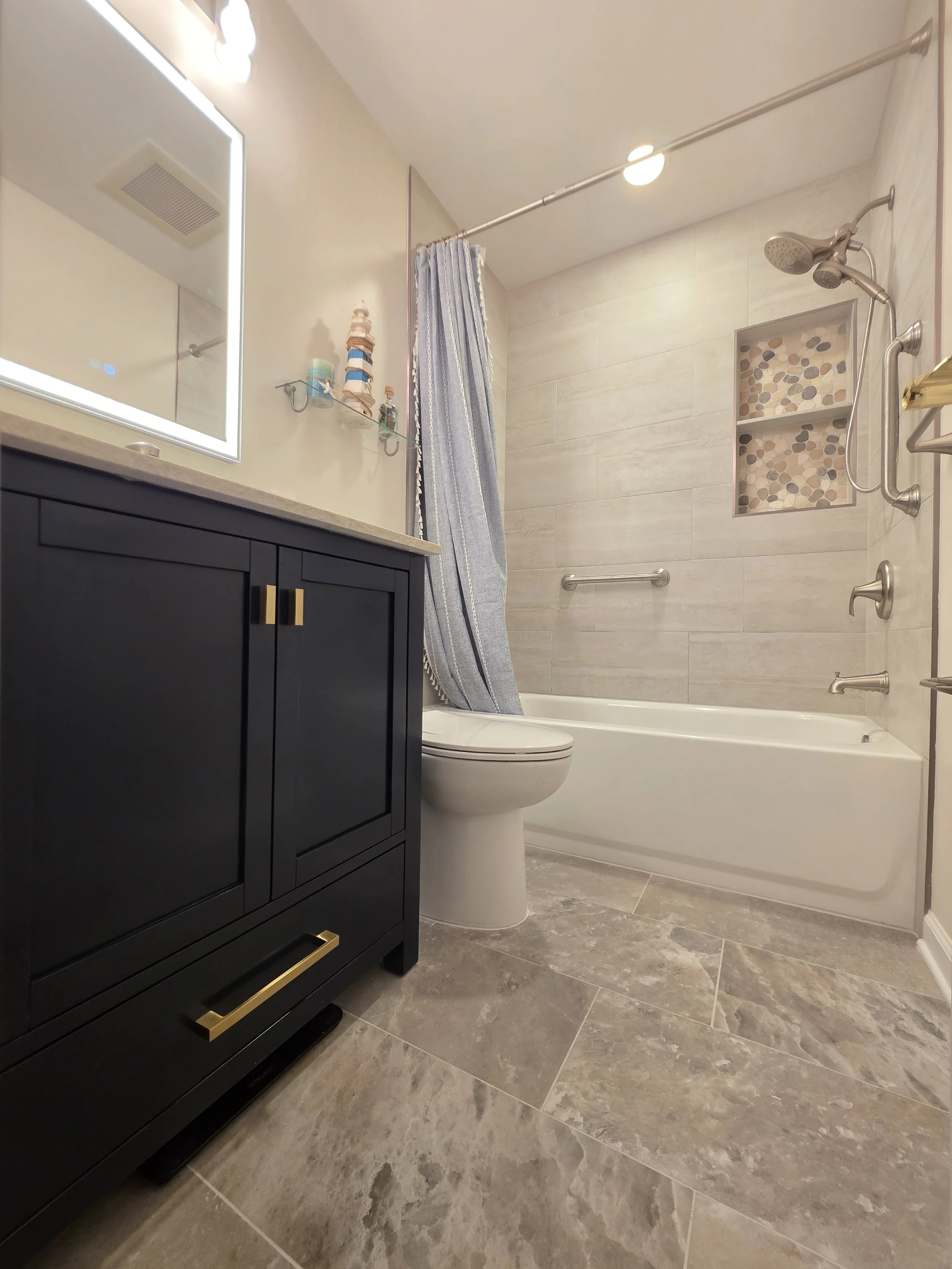 RoseAnn and Tom’s remodeled hallway bathroom with a navy vanity, illuminated mirror and tiled bathtub surround.