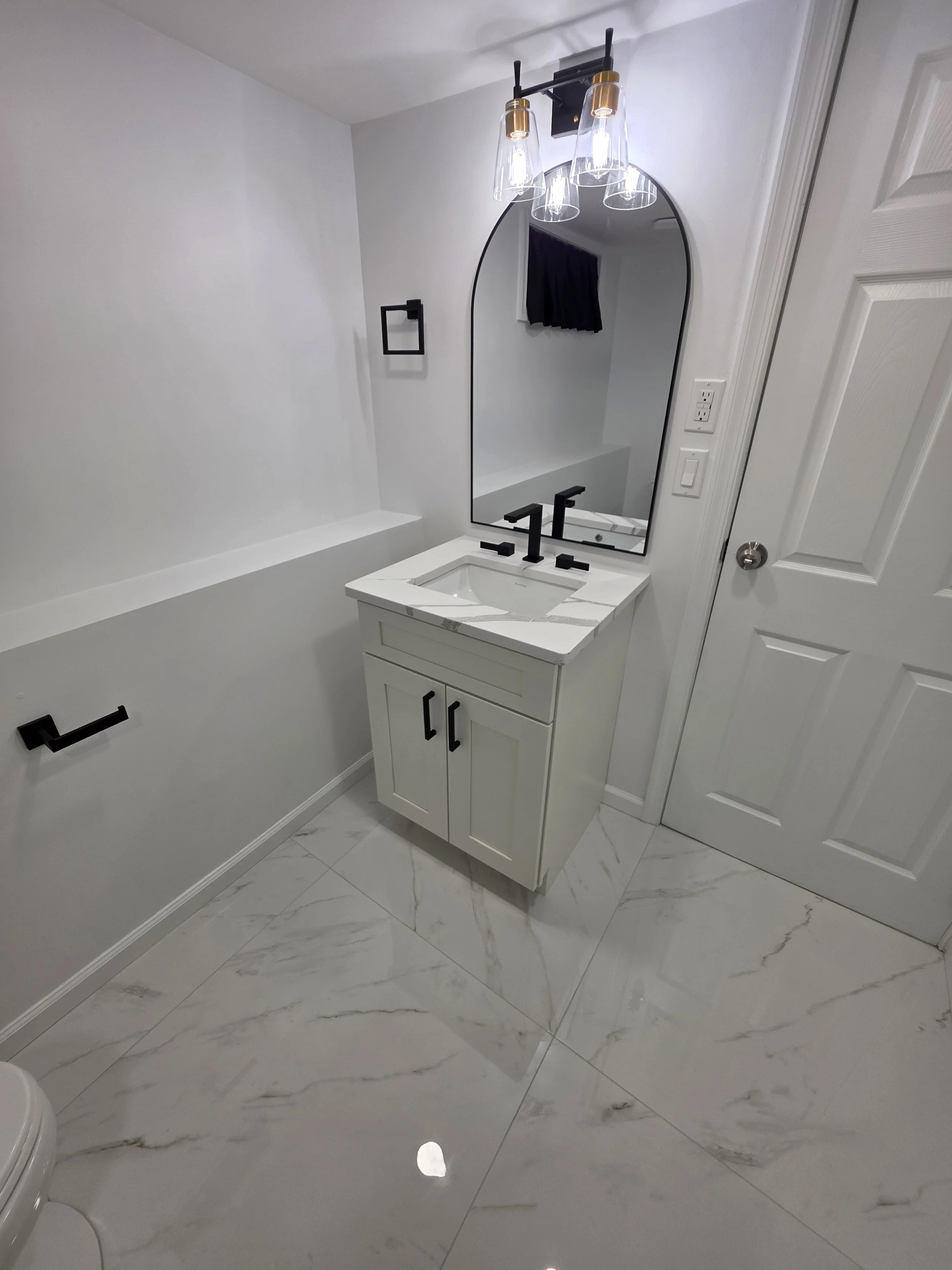 White vanity with black faucet, arched mirror and black cabinet hardware beside the bathroom door.