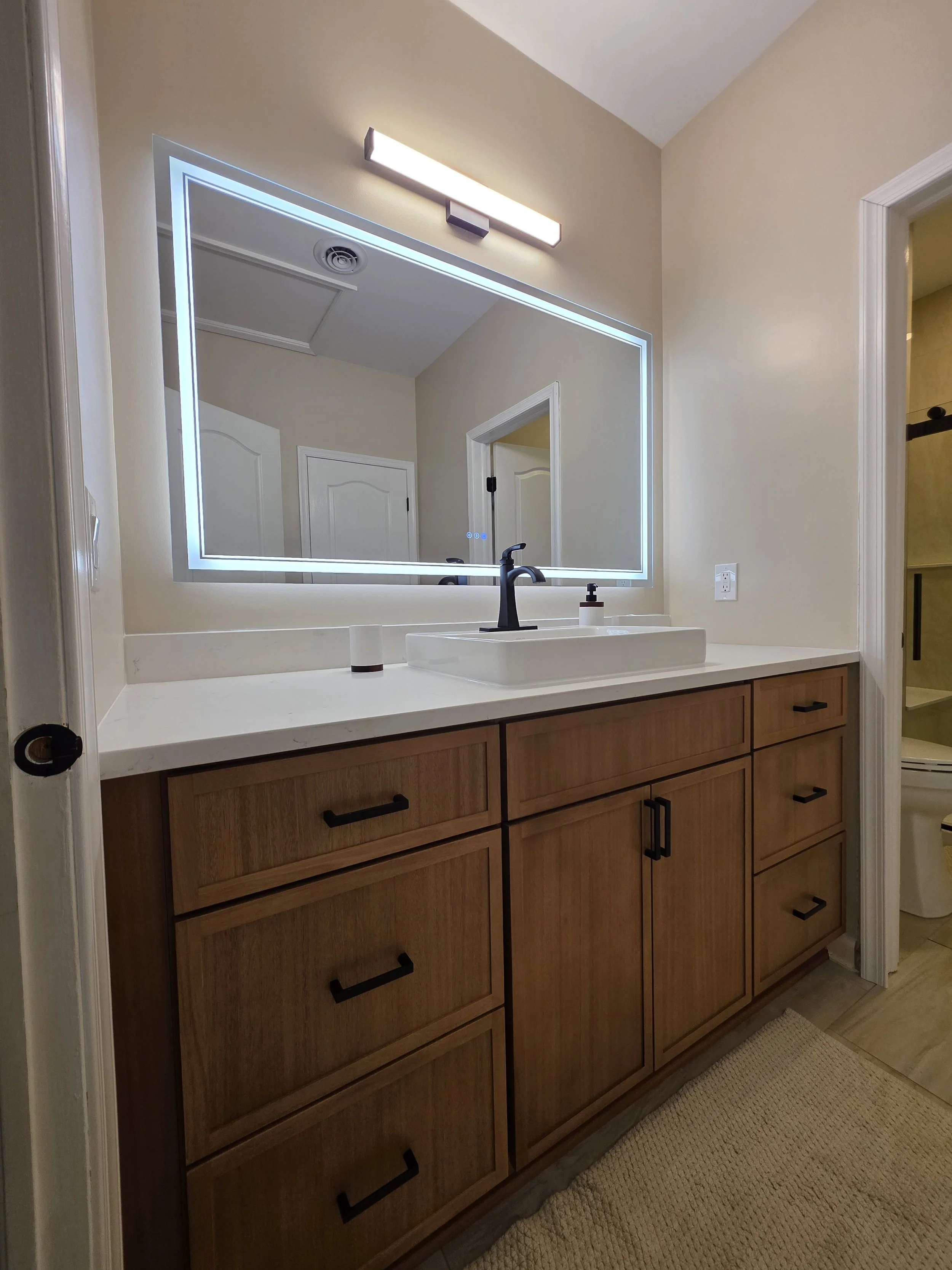 Wood-tone vanity with black hardware, a white countertop, raised rectangular sink and illuminated mirror.