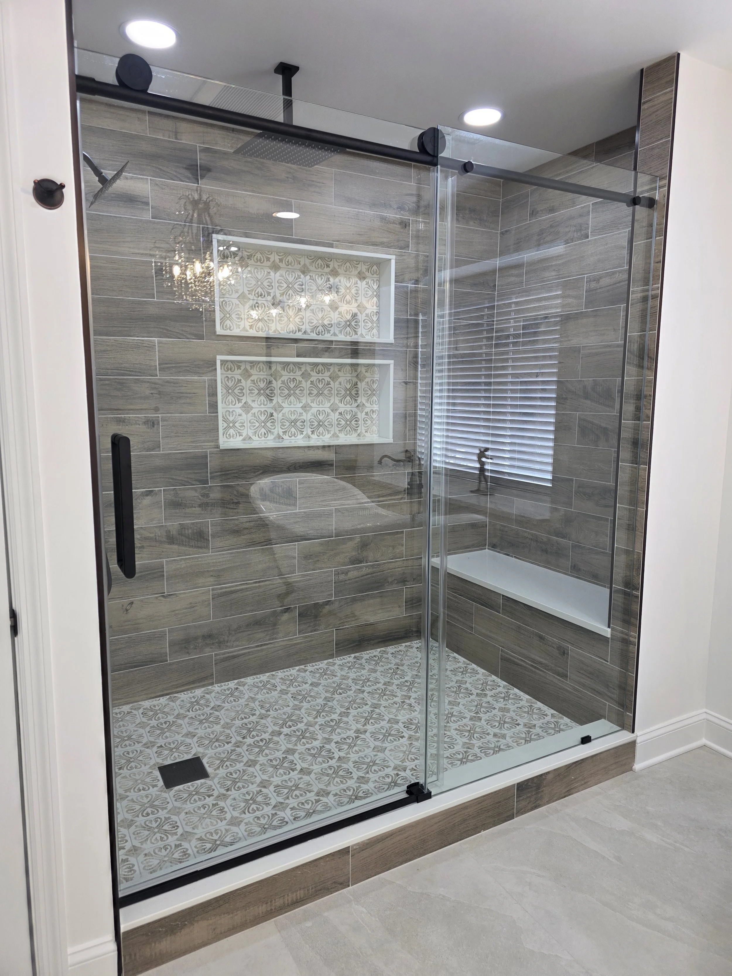 Glass shower enclosure with black sliding hardware, wood-look wall tile and two patterned recessed niches.