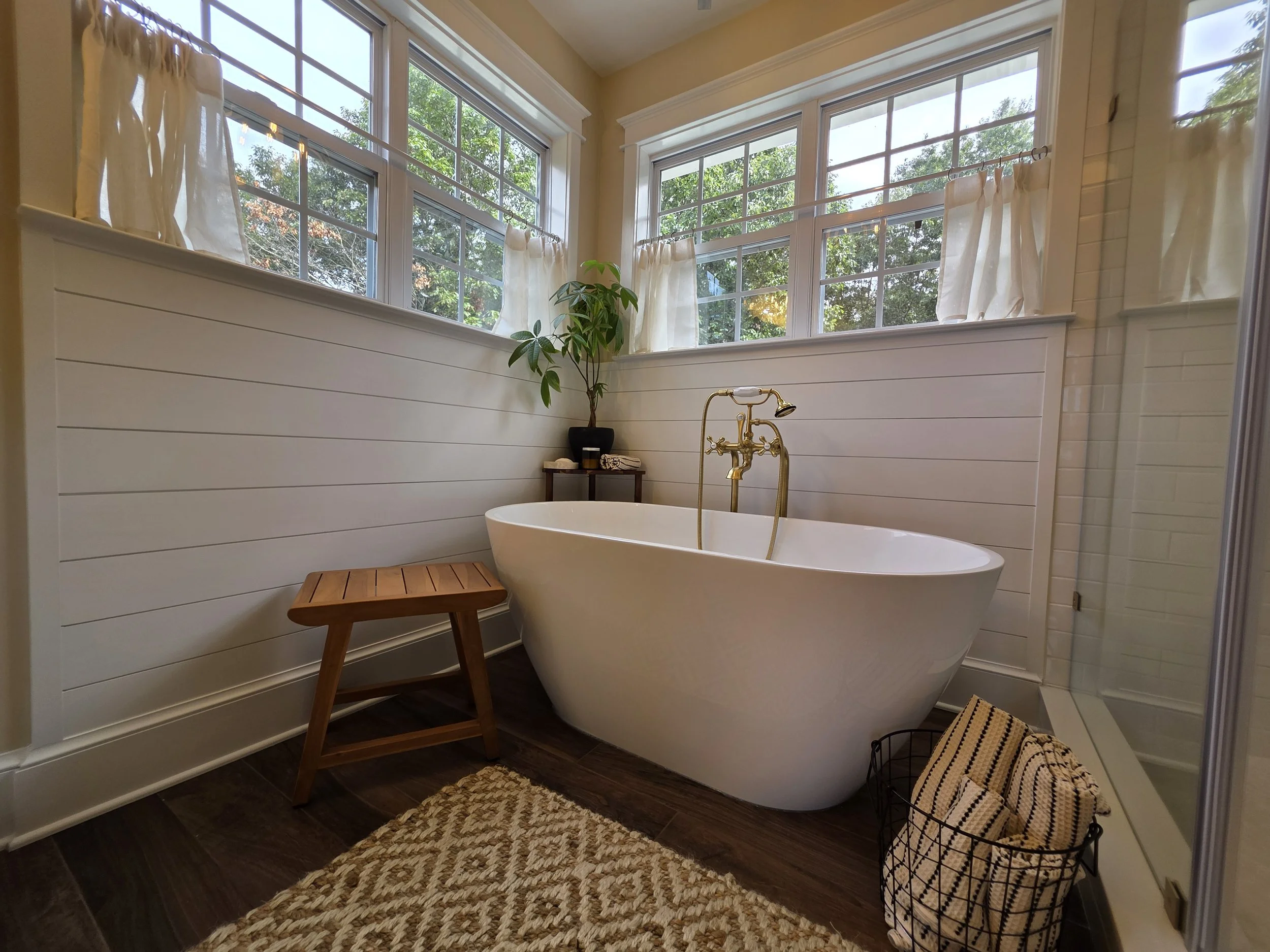 Freestanding tub beneath corner windows with wall paneling.