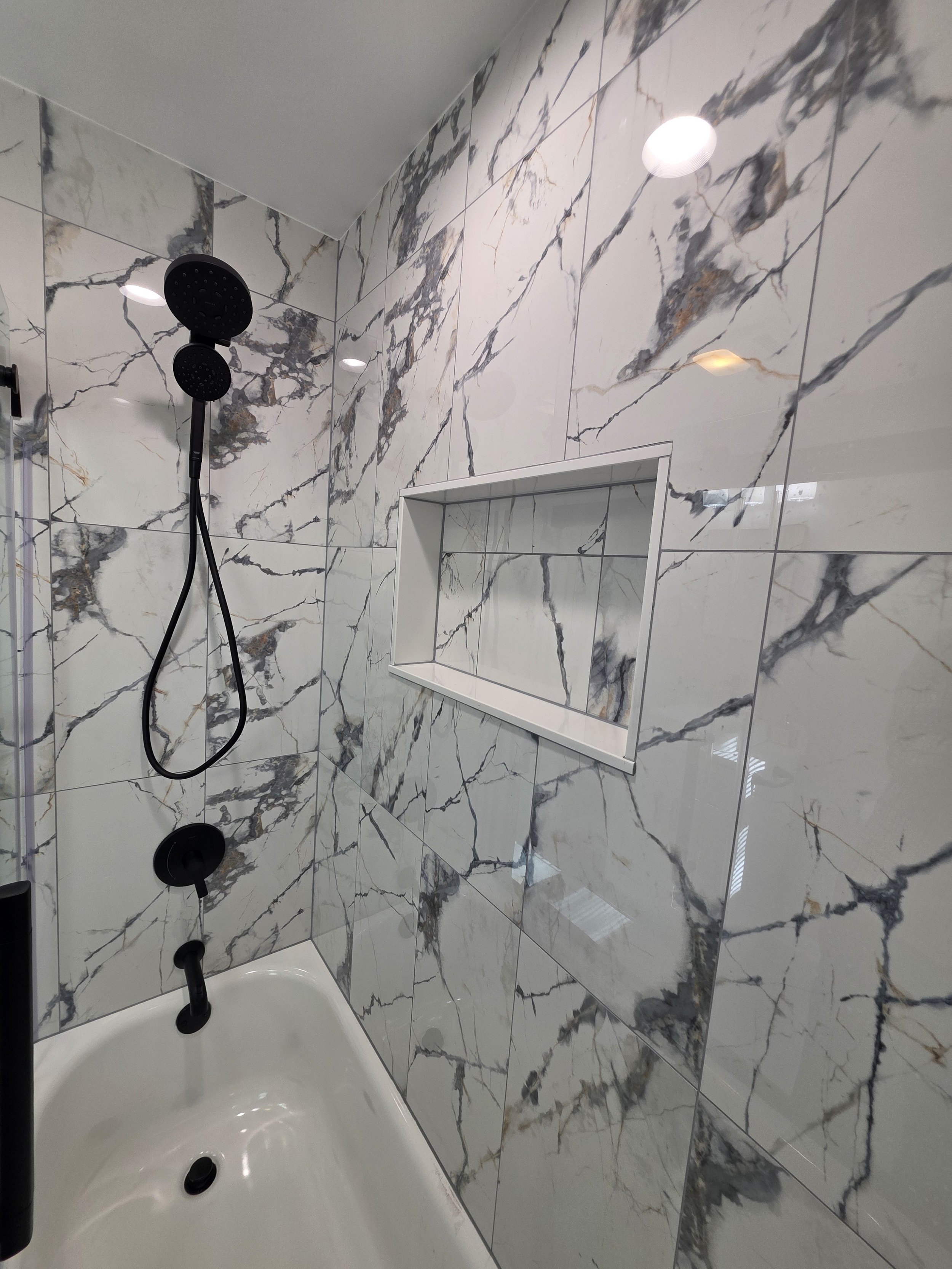 Black handheld shower and tub fixtures beside the recessed niche in the veined tile surround.