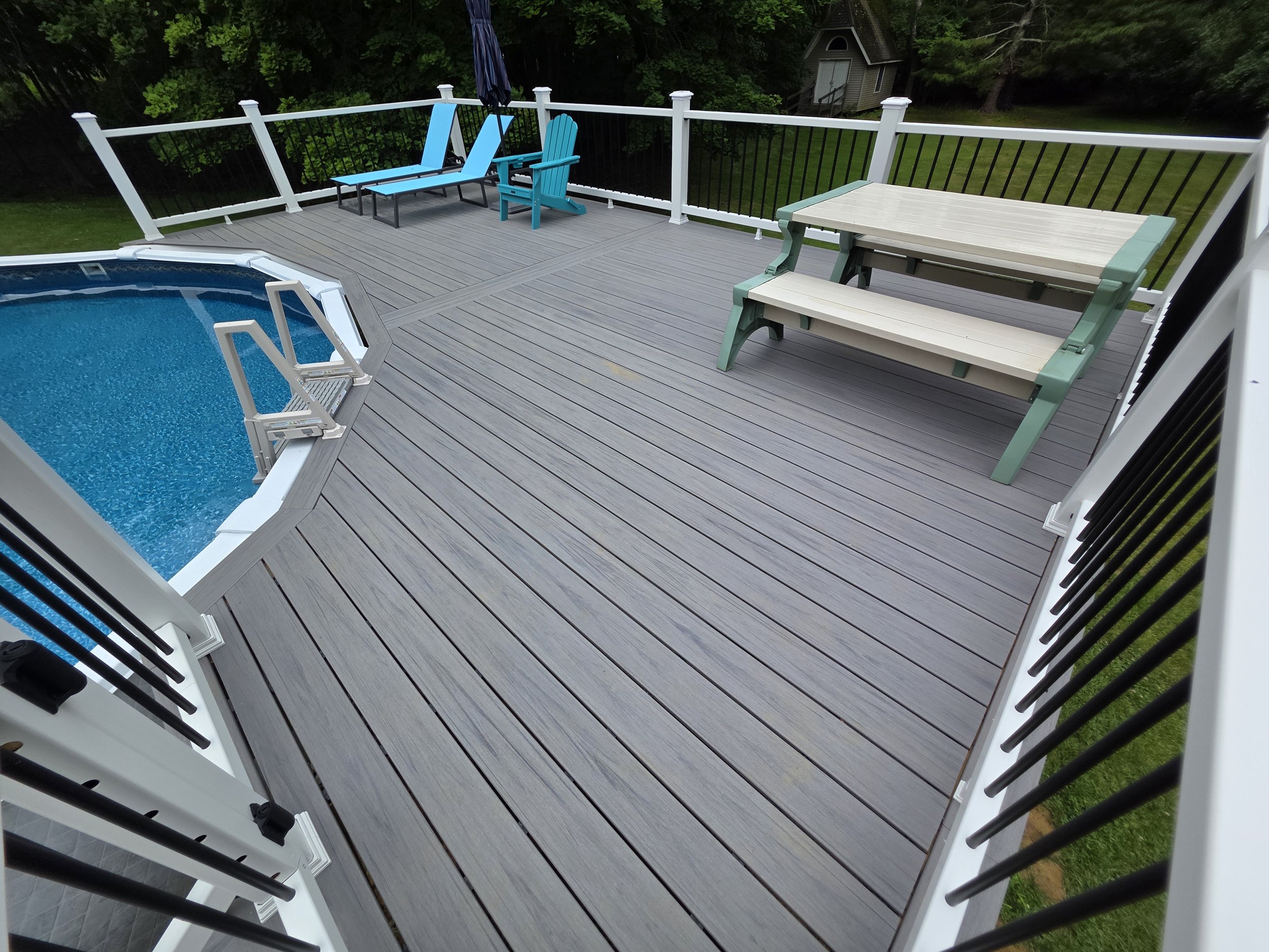 Gray pool deck with a picnic table and blue chairs, enclosed by white railings with dark balusters.