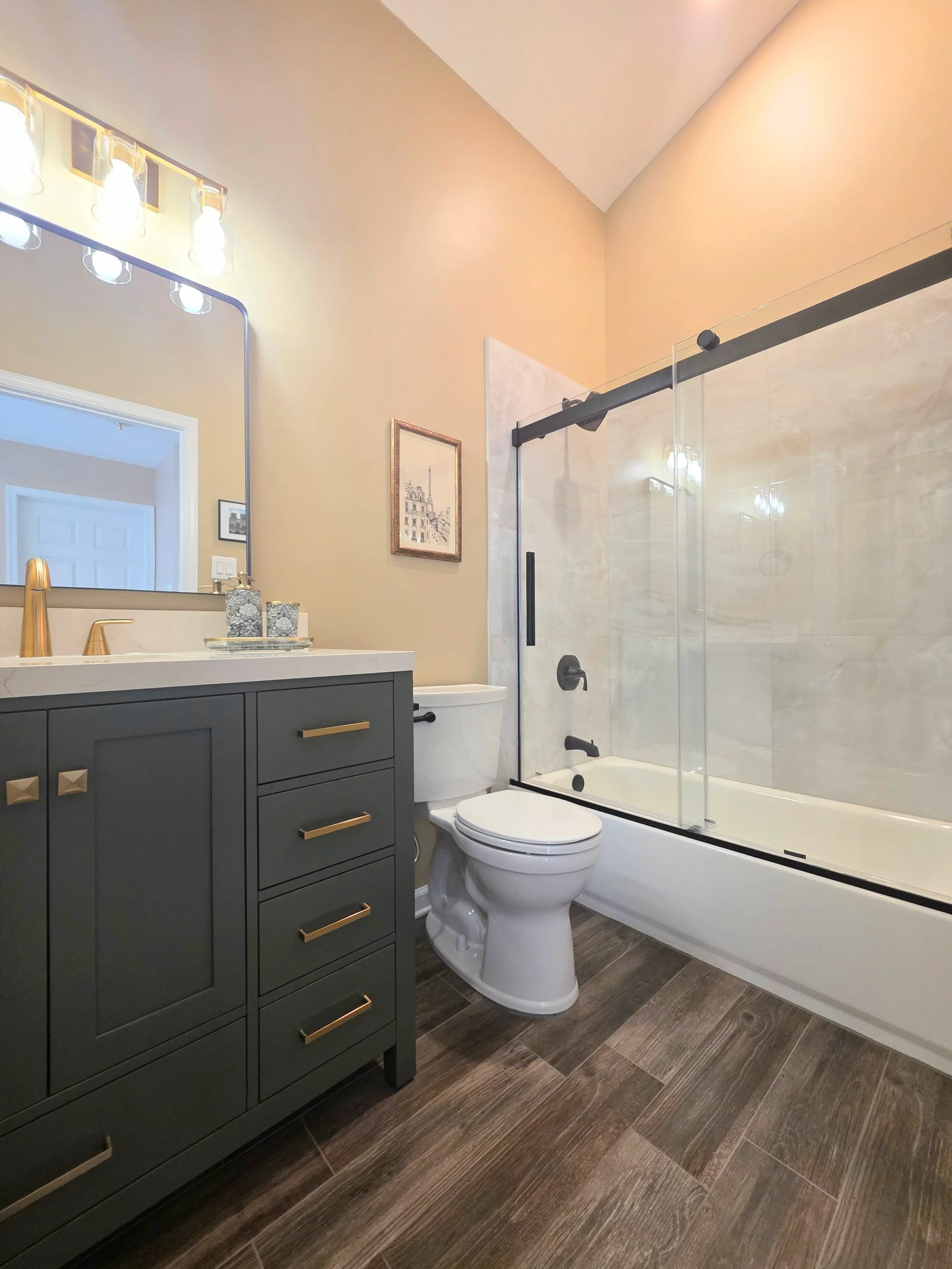 Joe and Maria’s remodeled hallway bathroom with a green vanity, glass tub enclosure and wood-look flooring.