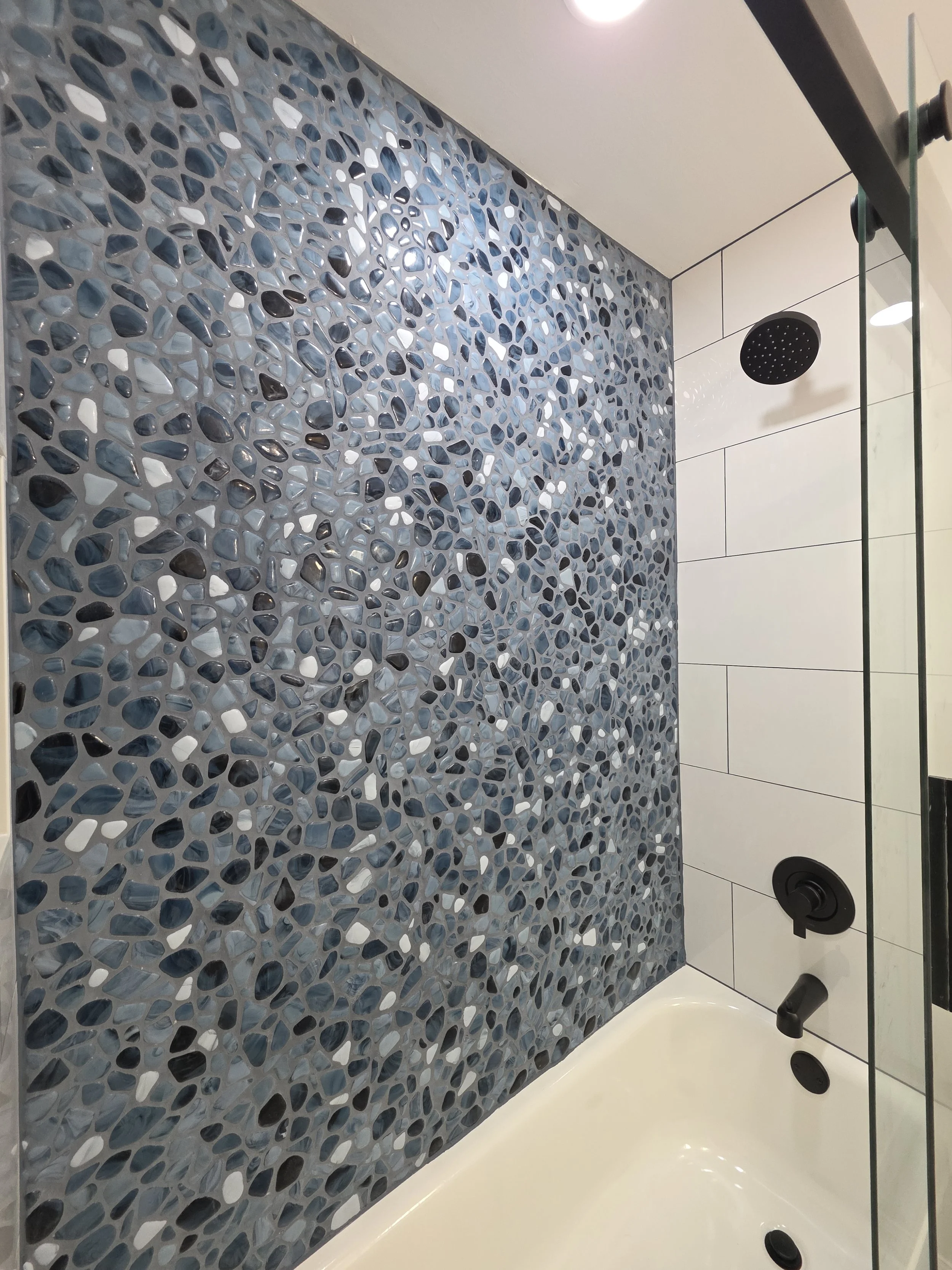Blue river rock accent tile above the white bathtub, with black shower fixtures on the adjoining white-tile wall.
