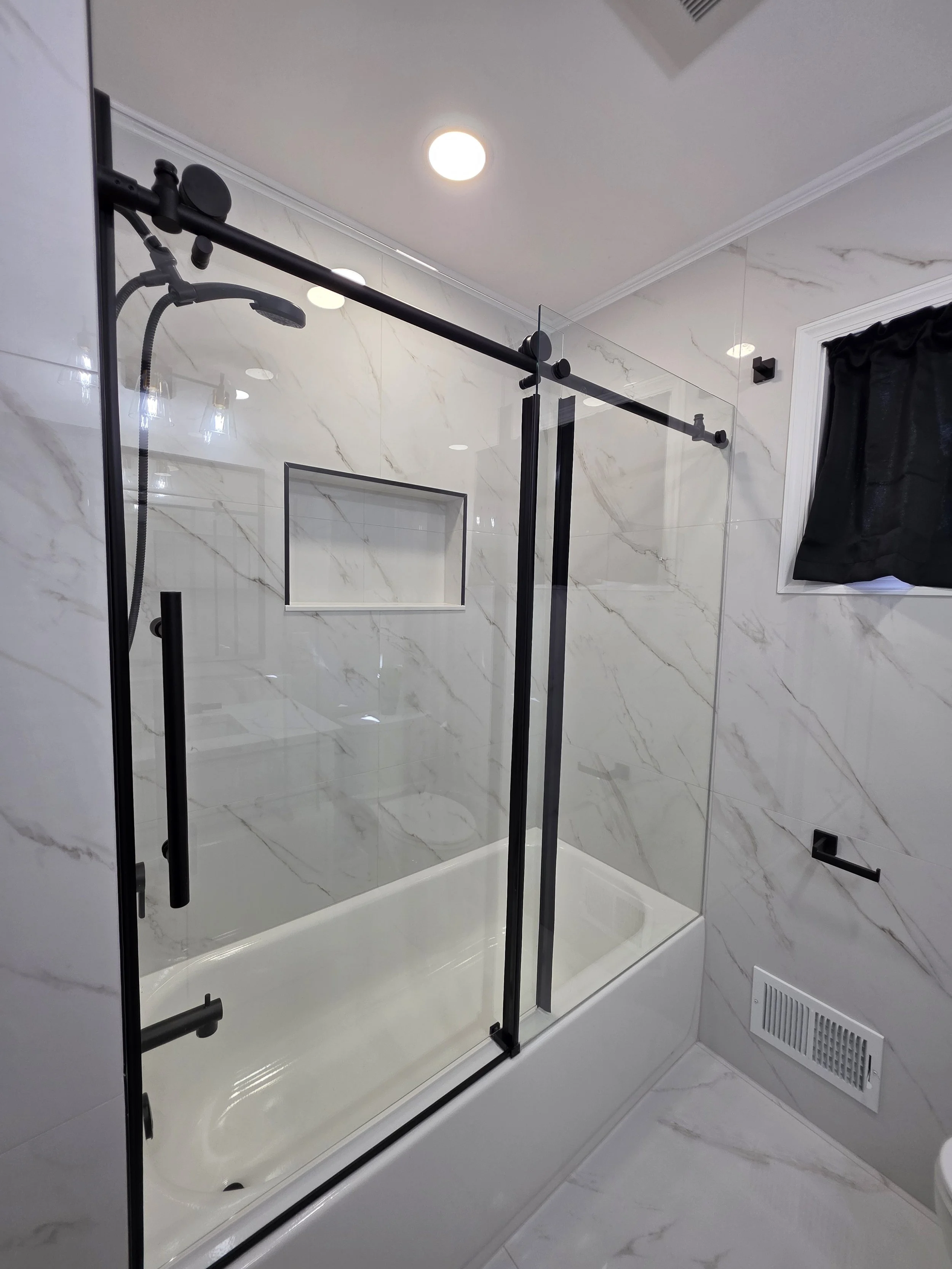 Glass tub enclosure with black sliding-door hardware, marble-look wall tile and a recessed shower niche.