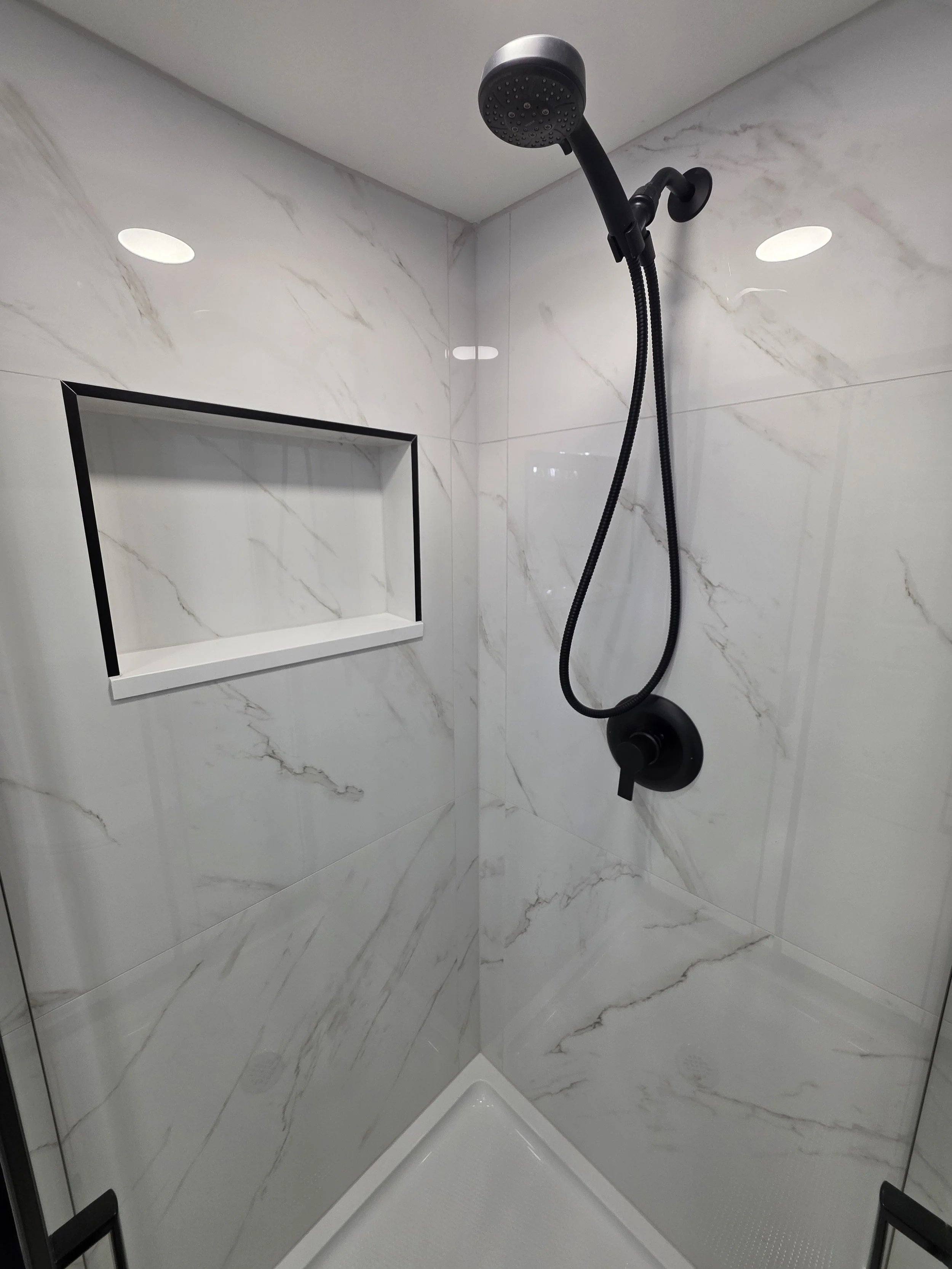 Black handheld shower and controls beside a rectangular niche in the marble-look shower wall.