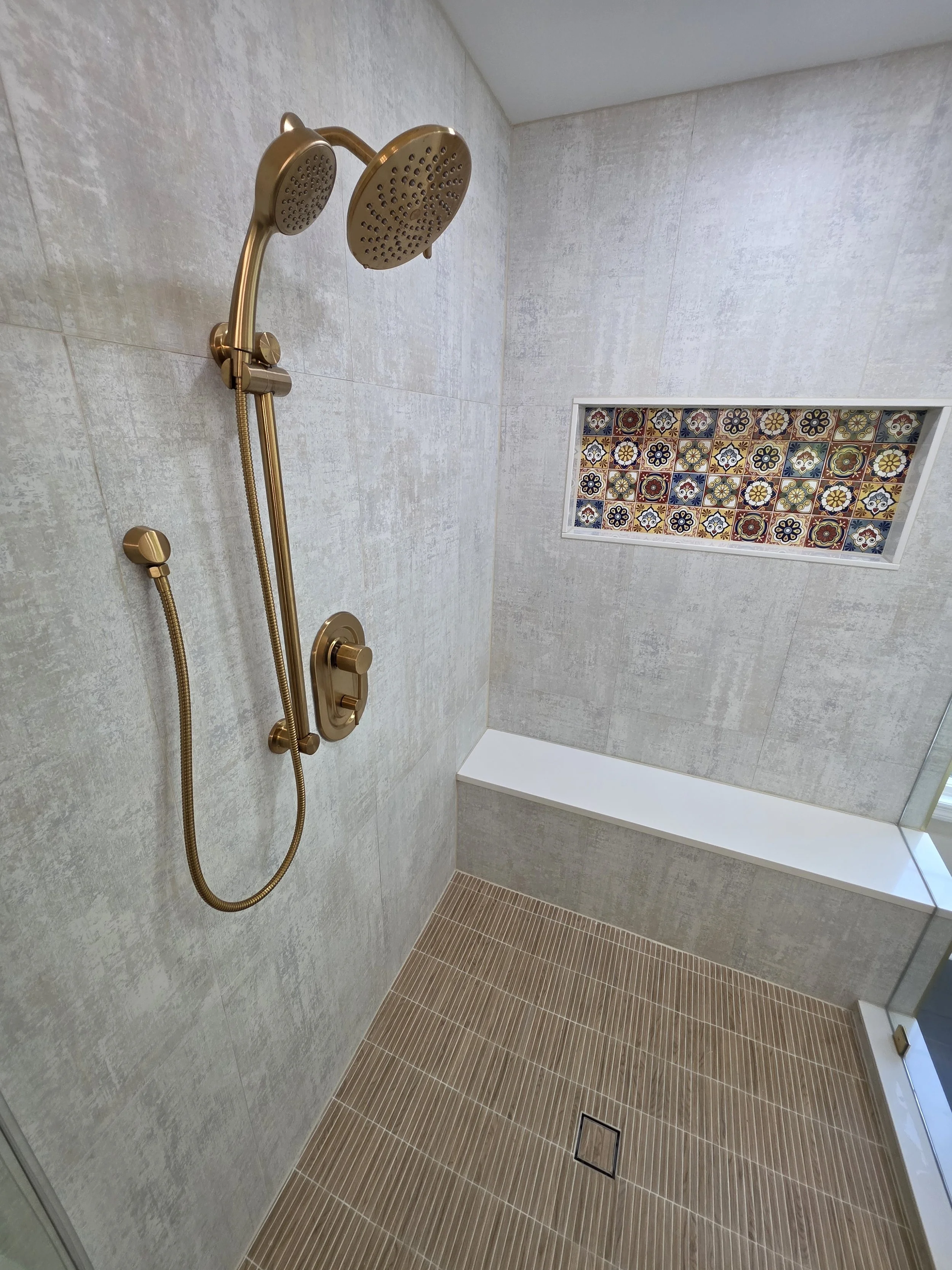 Gold-tone shower controls and handheld shower beside the patterned recessed niche and bench.