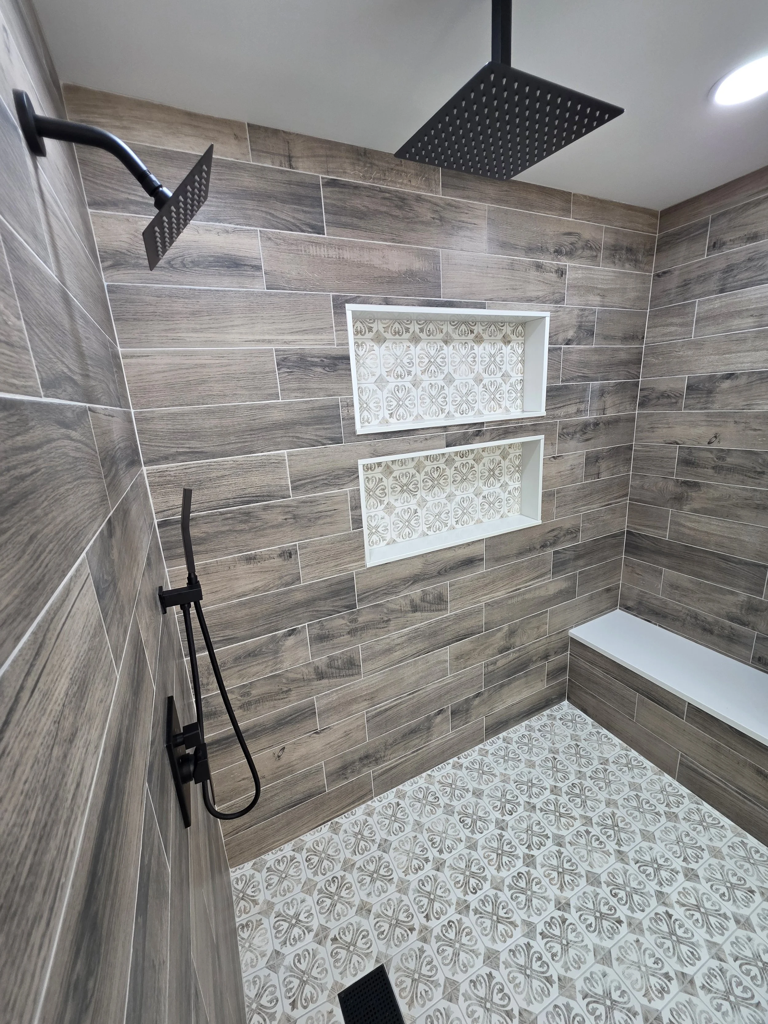 Shower interior with a rain showerhead, handheld spray, bench and patterned tile floor.