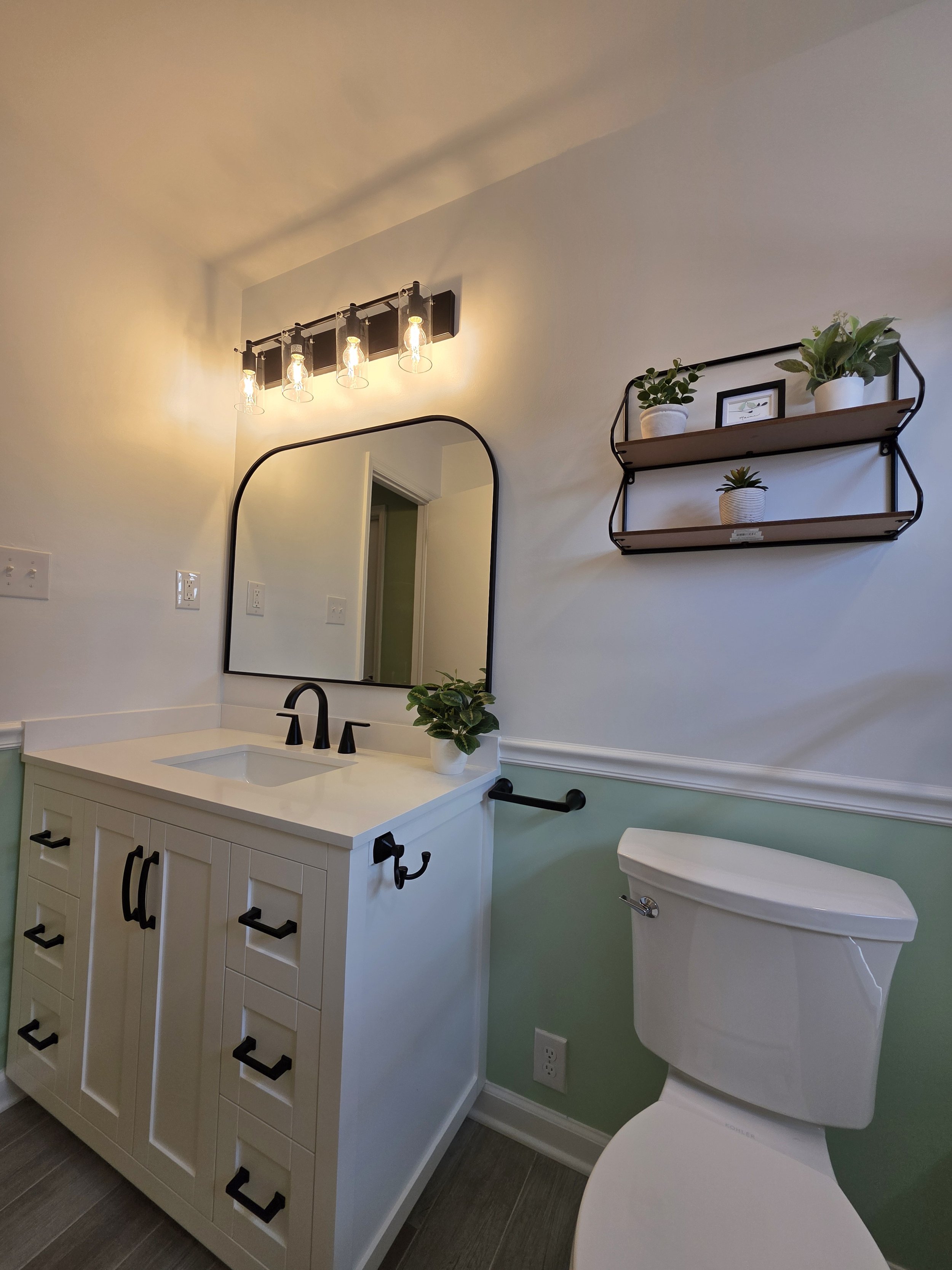 Vanity and toilet against a green lower wall, with a rounded mirror and two wall shelves.