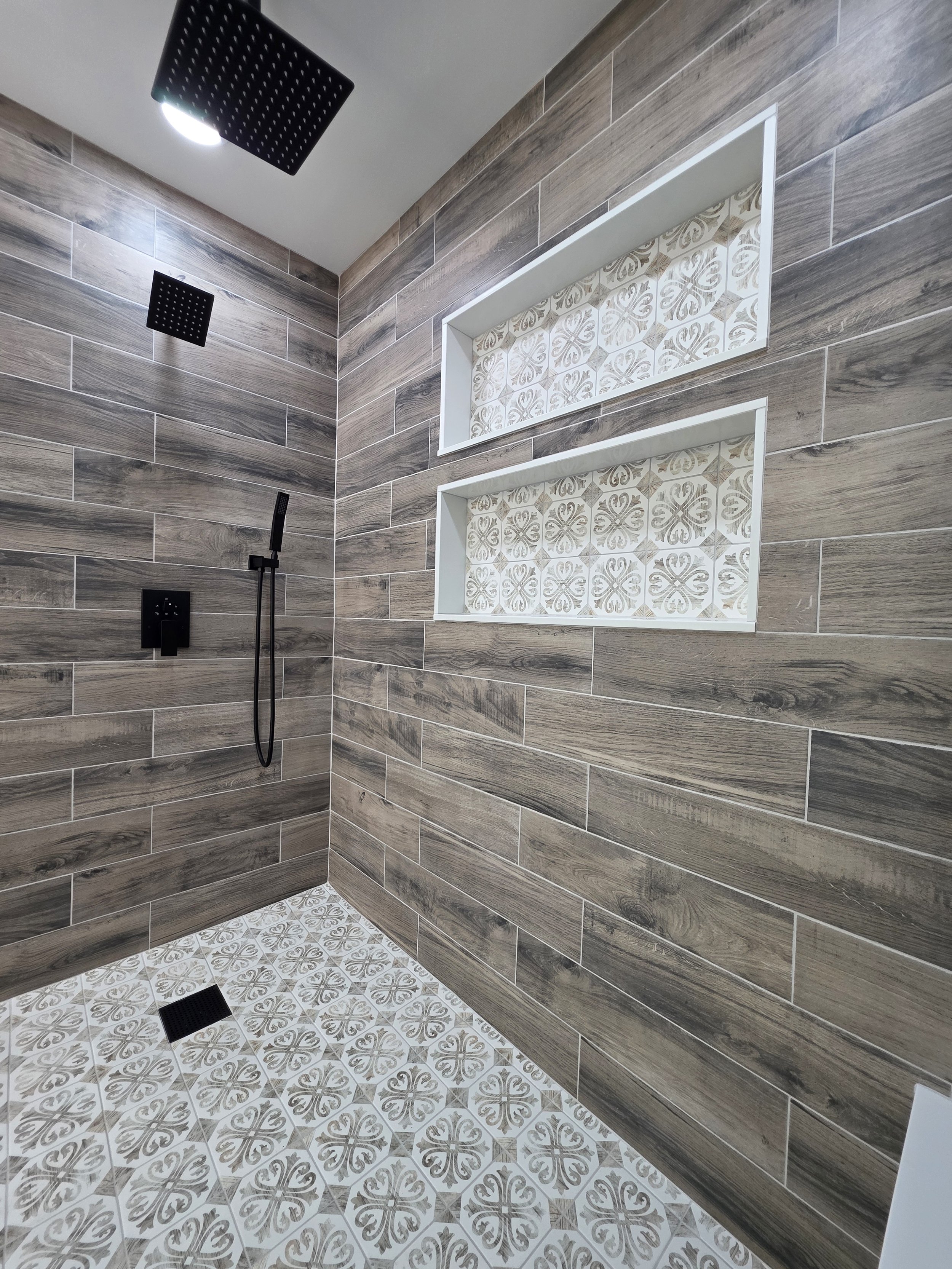 Wood-look shower tile with two stacked patterned niches, black fixtures and a patterned floor.