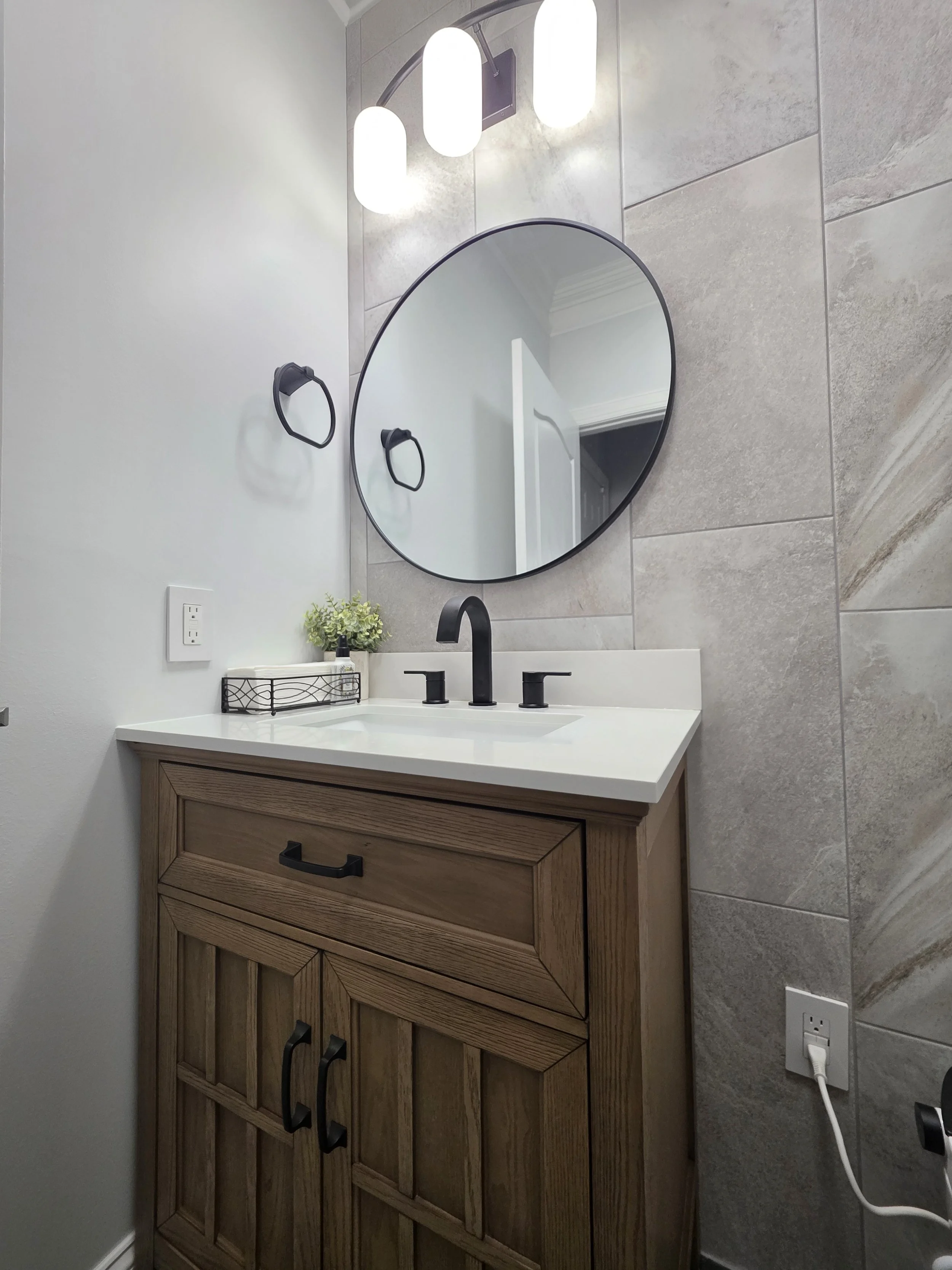 Close view of the wood-tone vanity, black faucet, round mirror and overhead lights.