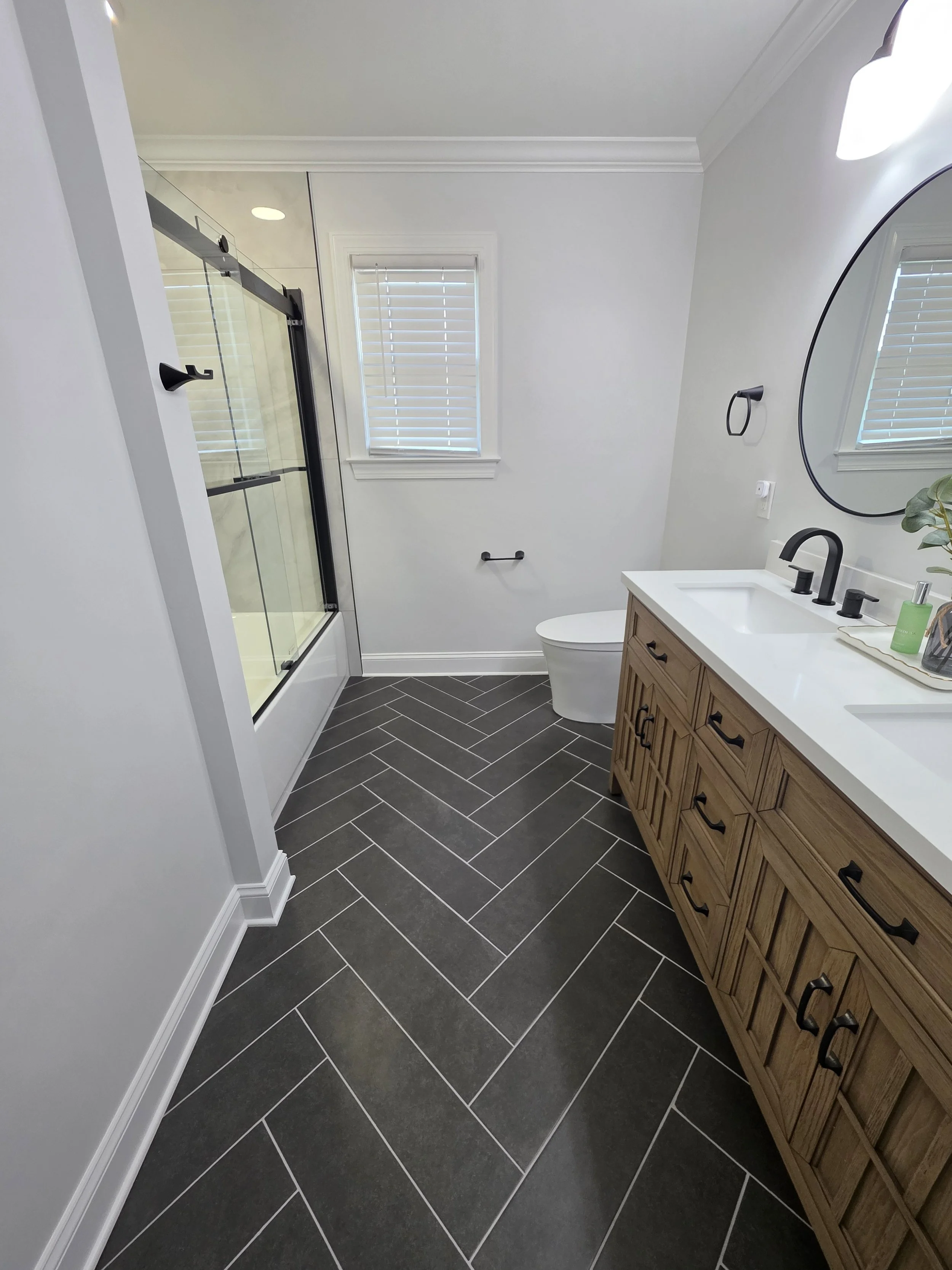 Finished hallway bathroom with a wood-tone double vanity, dark herringbone tile floor and glass tub enclosure.