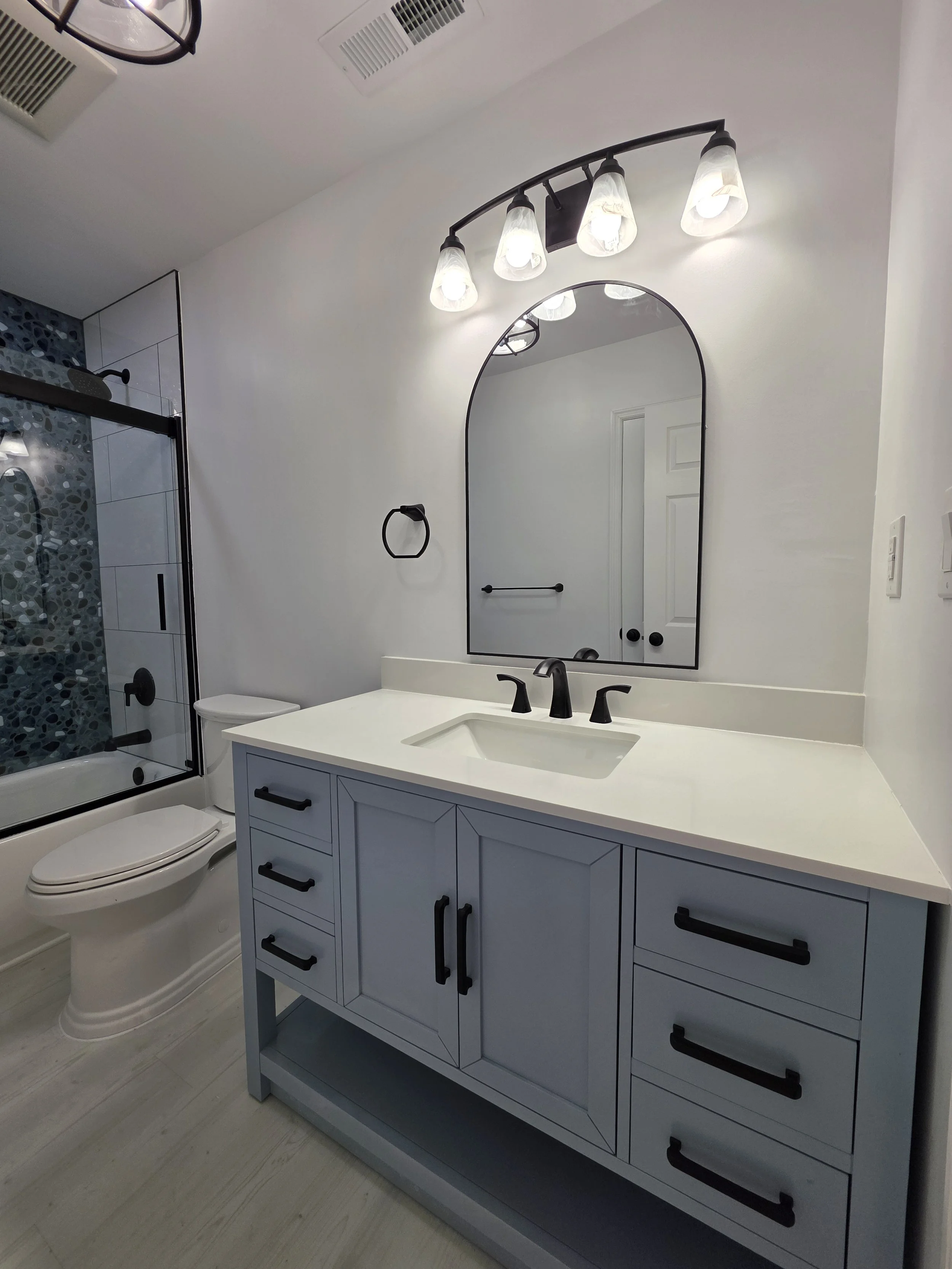 Gray vanity with a white countertop, black faucet and arched mirror beneath a three-light fixture.