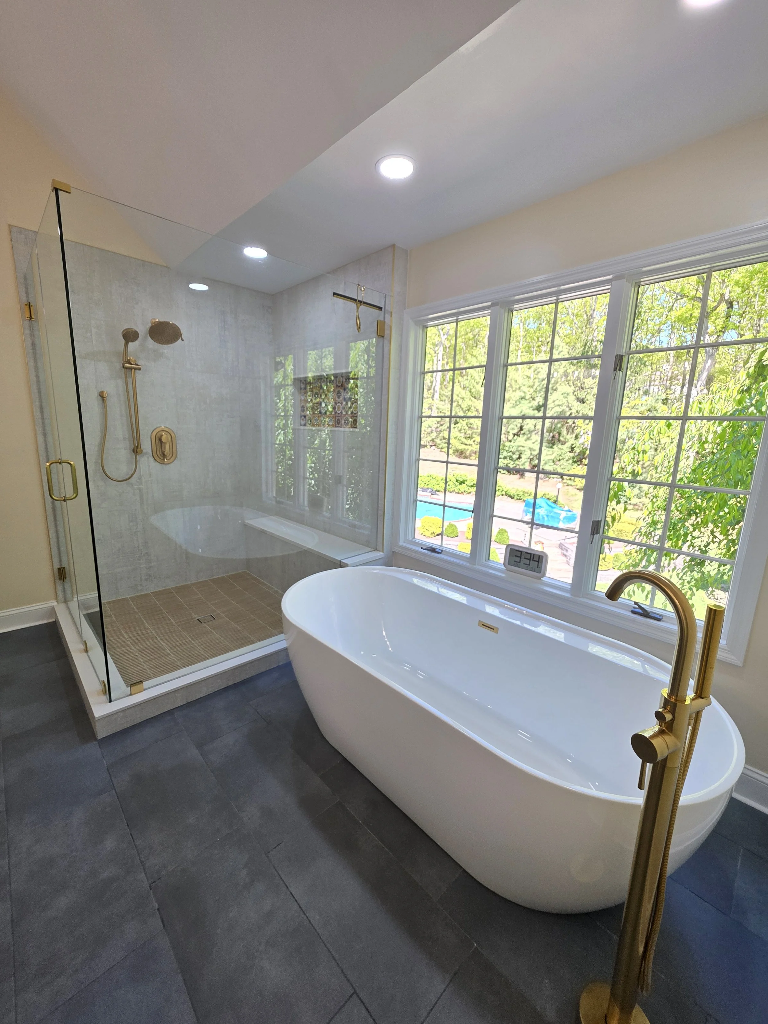 Freestanding white tub beside a large window, with gold-tone tub filler and glass shower in Ricardo and Christine’s master bathroom.